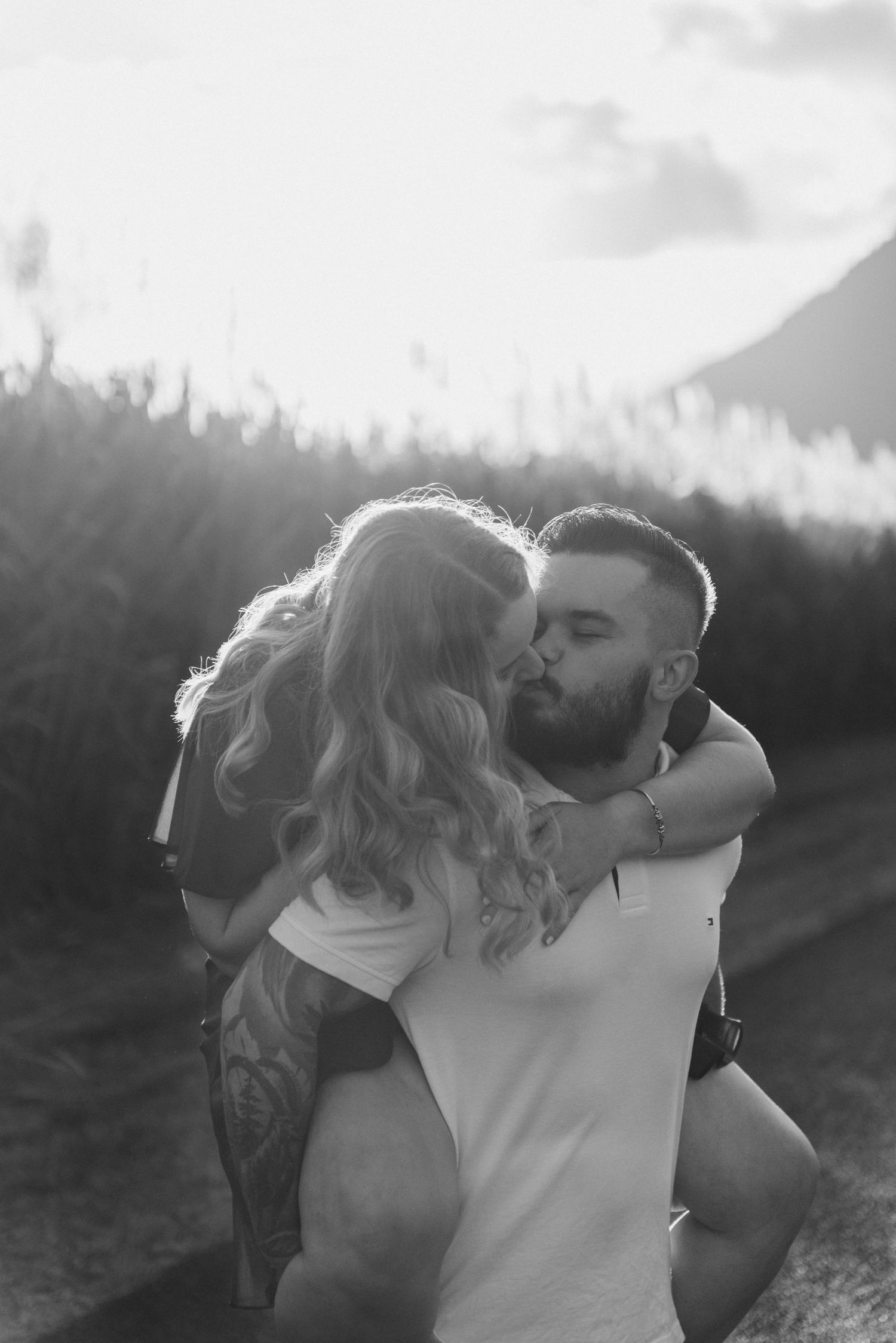 Black and white photo of a couple sharing a romantic kiss outdoors at sunset.