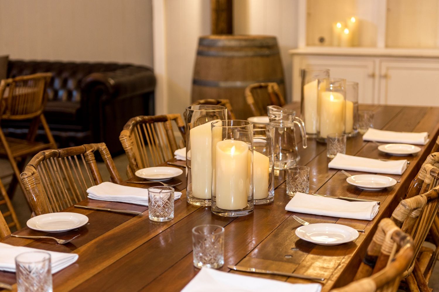 Intimate rustic dining table set with pillar candles, wooden chairs, white plates, and a wine barrel backdrop.