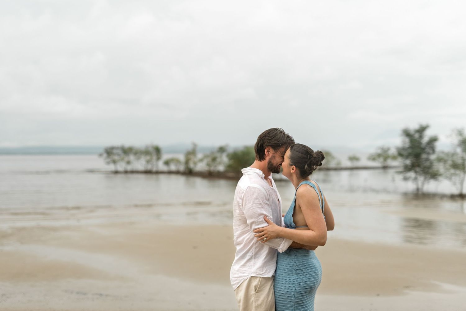 Couple embracing and kissing on a serene beach with calm water and mangroves behind them.