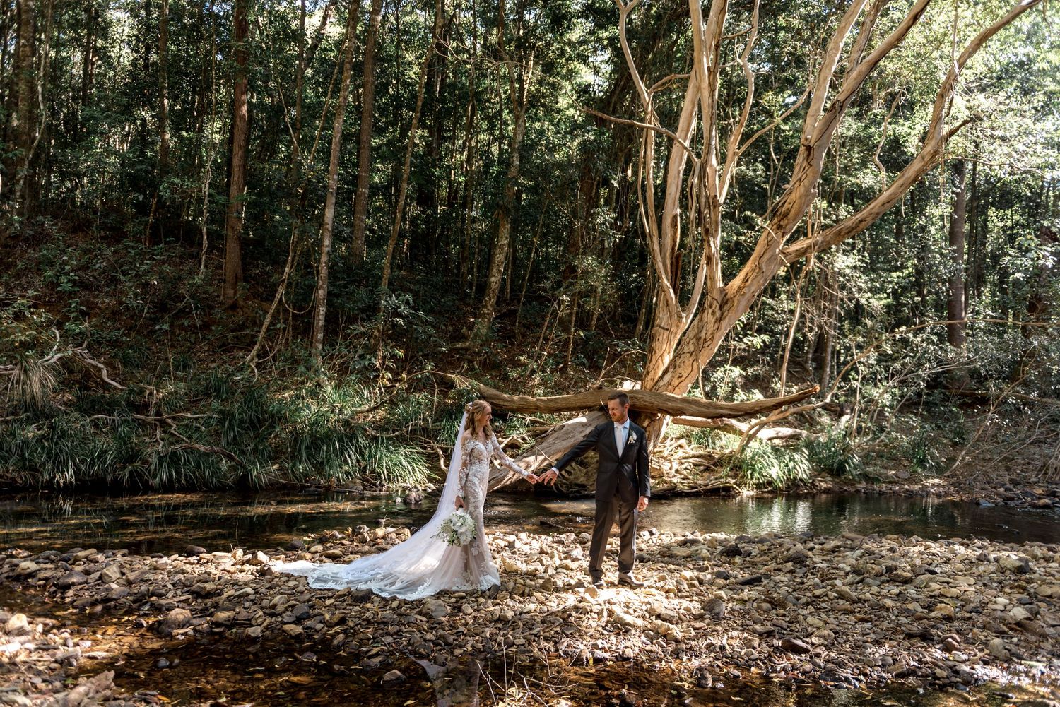 Bride and groom hold hands on rocky creek bed surrounded by tall forest trees.