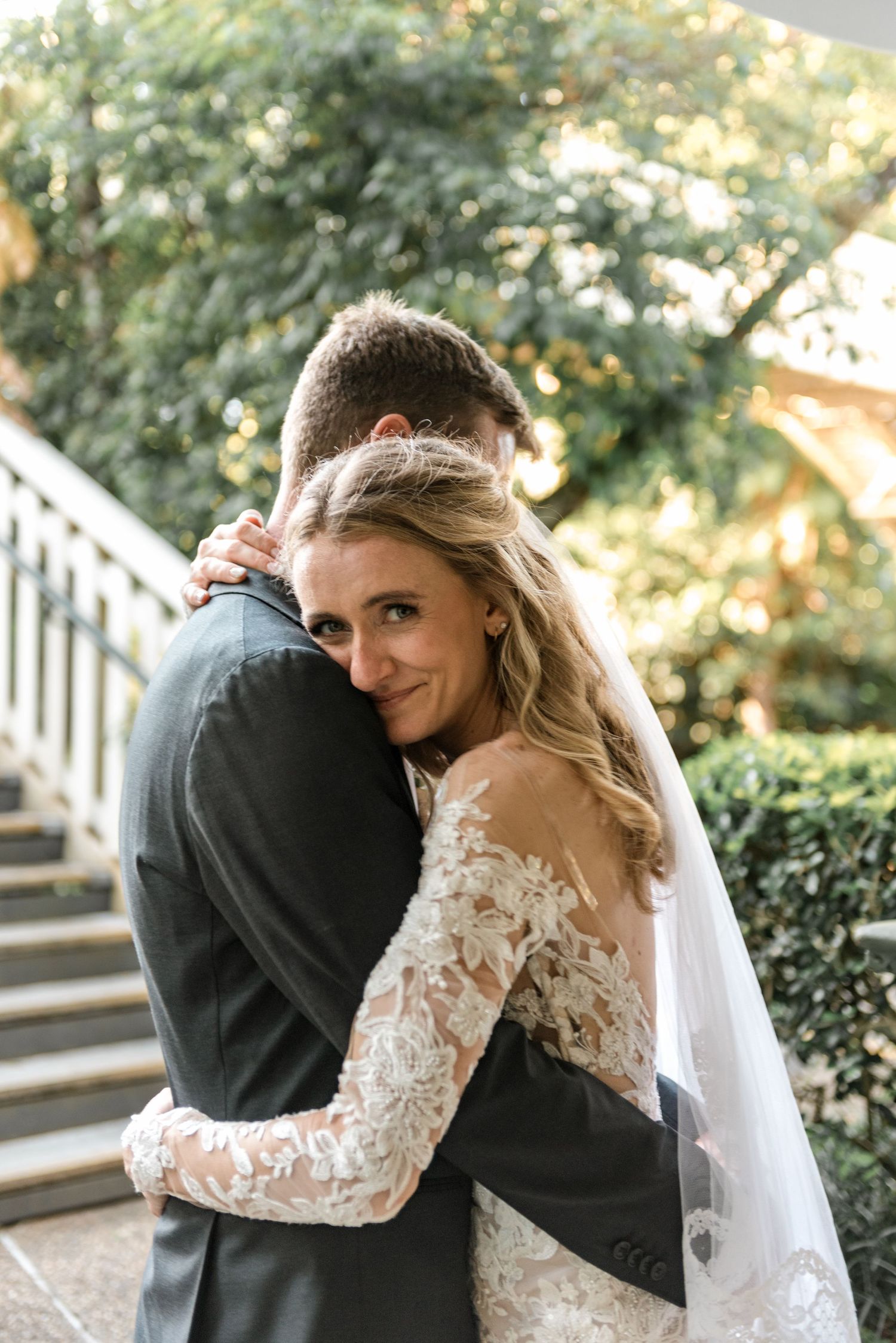 Smiling bride in lace long-sleeve gown embraces groom while looking back at camera in garden setting.