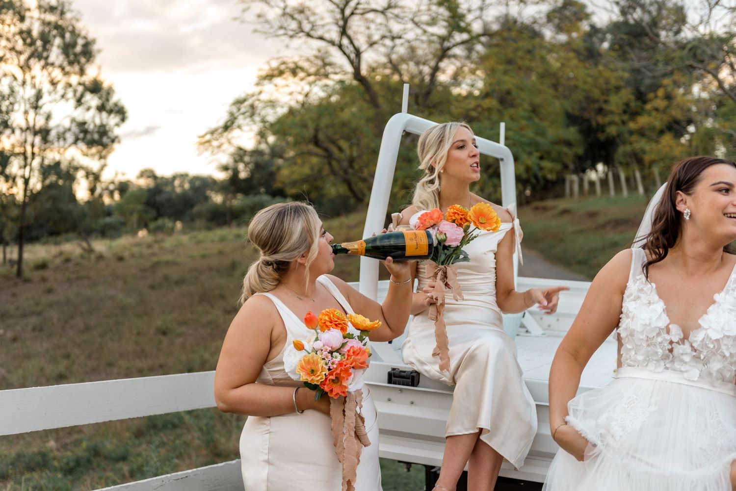 Bridesmaids in white dresses celebrate with champagne on a white truck tailgate at a rustic farm wedding.