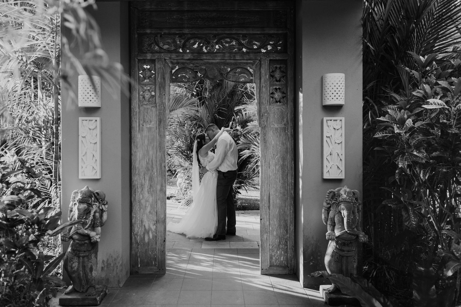 Black and white photo of bride and groom embracing in ornate Balinese carved doorway.