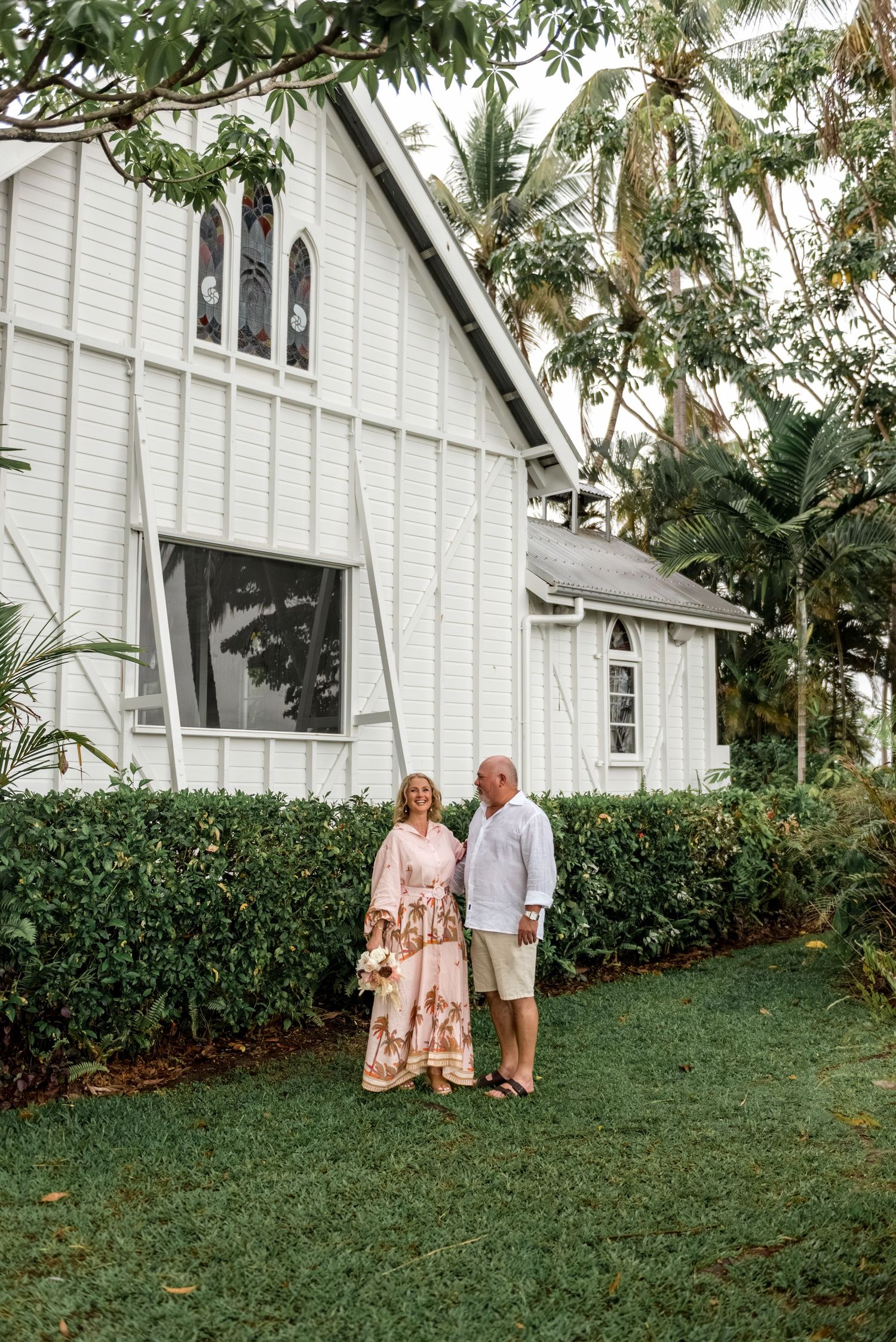 Couple standing together in front of a charming white heritage chapel with lush gardens.