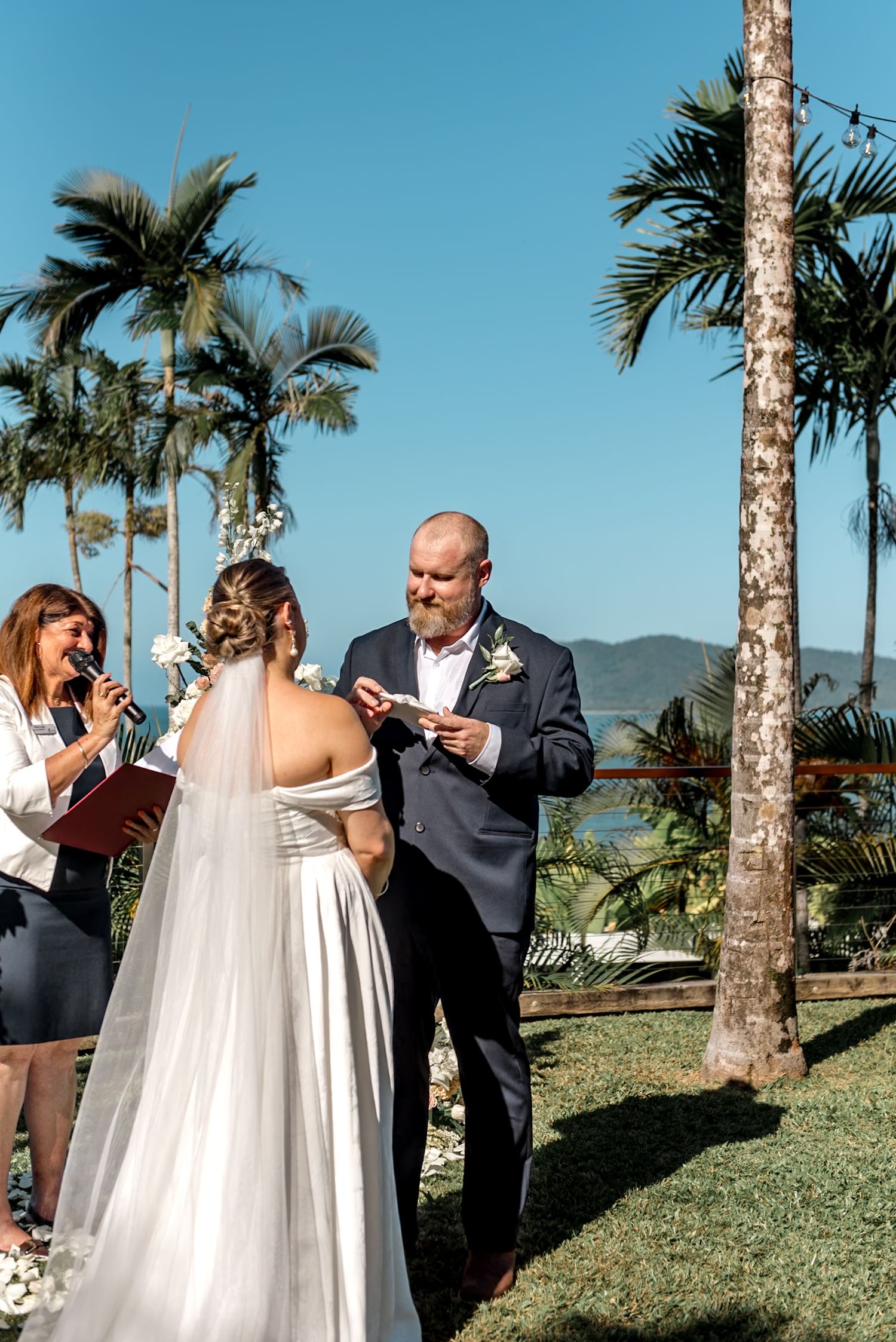 Groom places wedding ring on bride's finger during outdoor tropical ceremony with palm trees and blue sky.