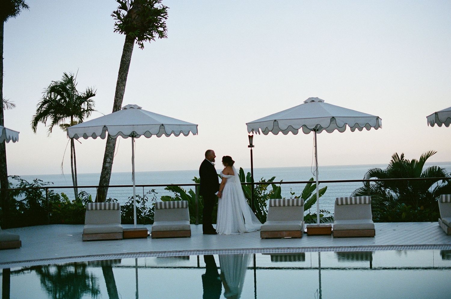Bride and groom silhouetted by a pool at dusk with white umbrellas and ocean views.