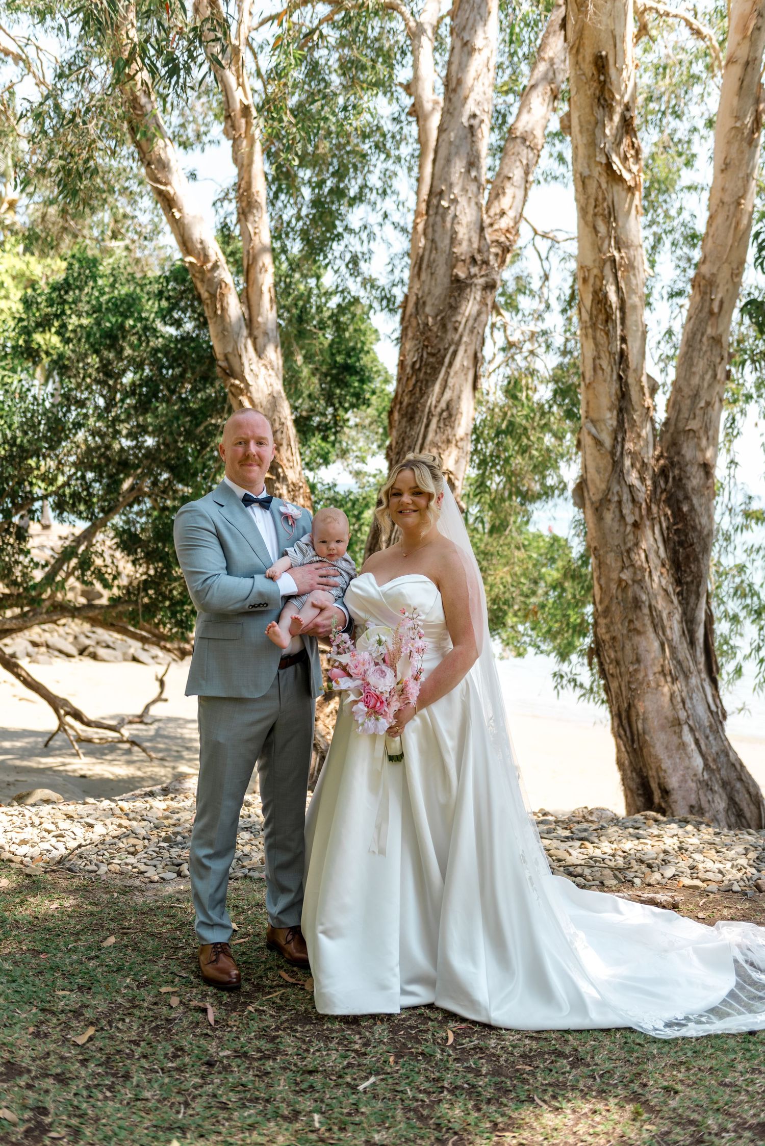 Newlywed couple holding baby in arms posing with pink bridal bouquet among tall eucalyptus trees outdoors.