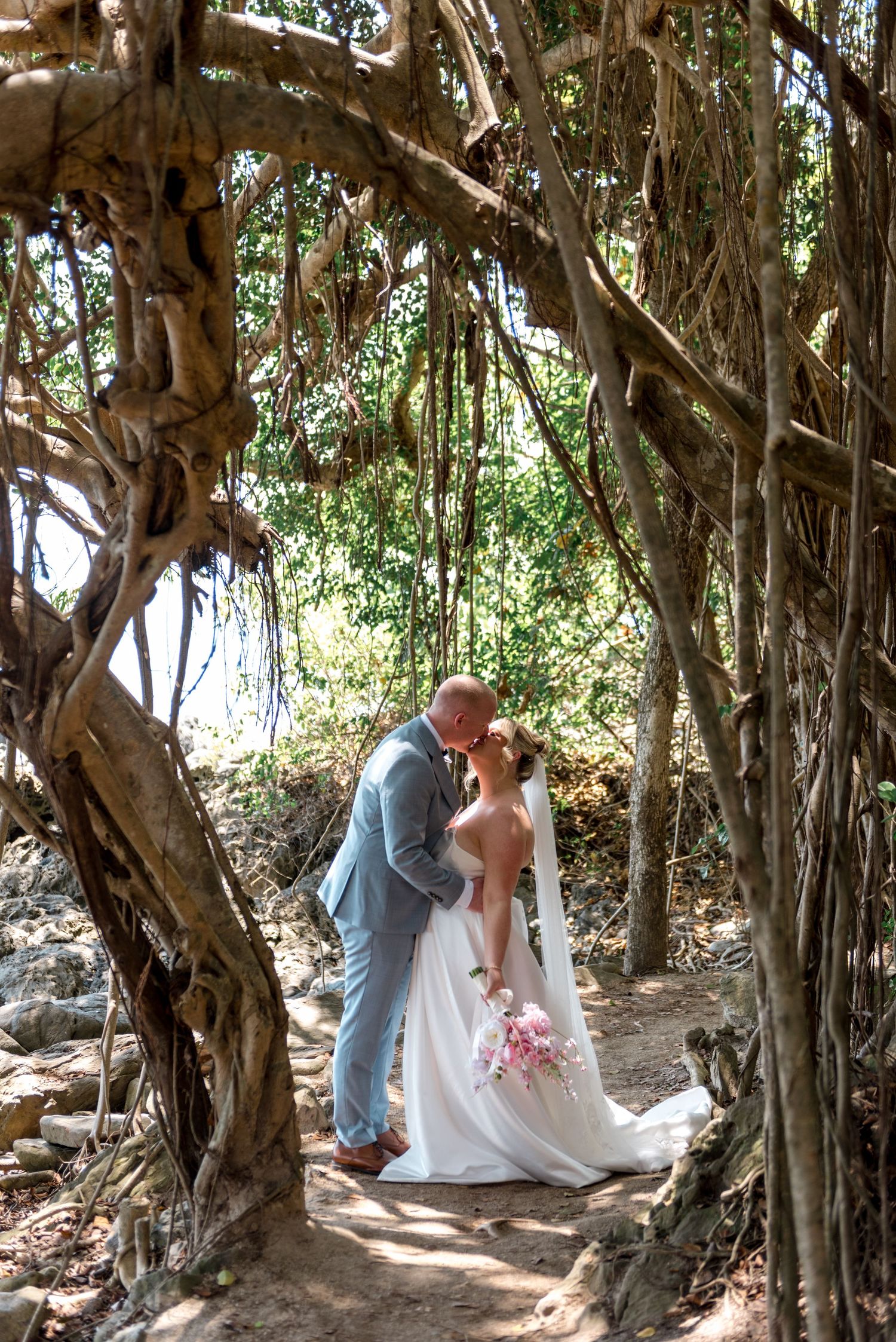 Couple shares a romantic kiss beneath dramatic twisted banyan tree roots on their tropical wedding day.
