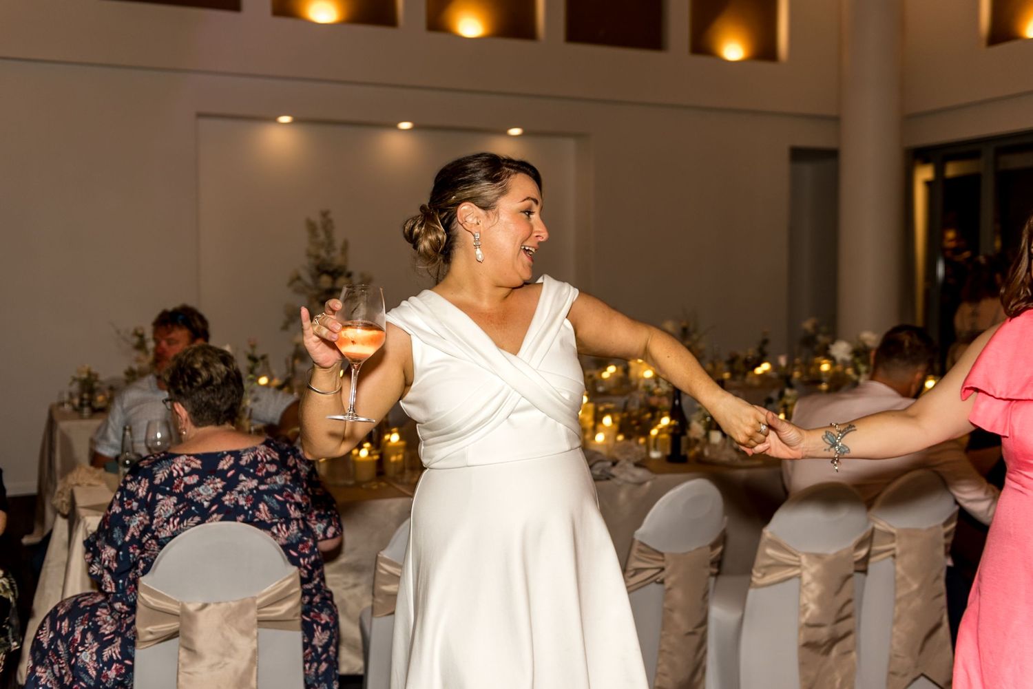 Bride in white dress laughs while holding a wine glass on the dance floor.