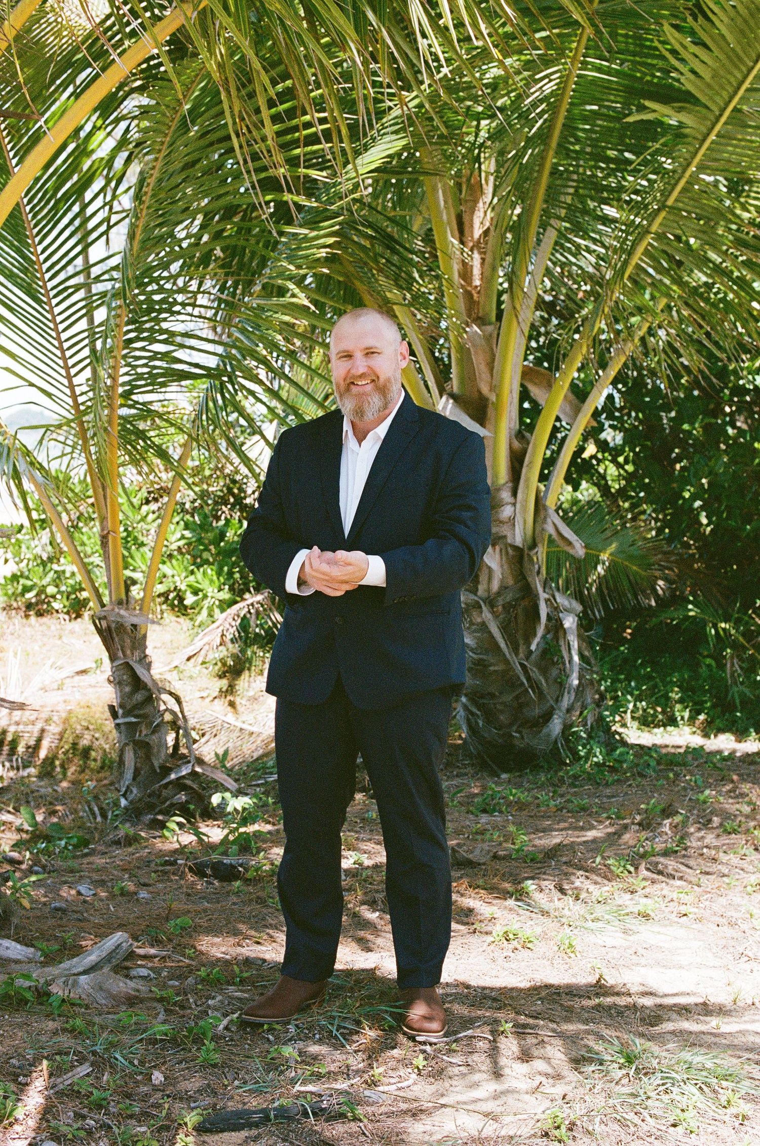 Smiling groom in navy suit stands under tropical palm trees on sunny wedding day.