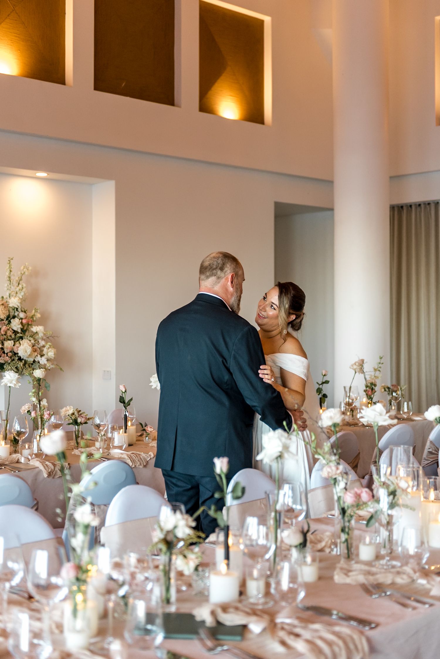 Newlyweds share an intimate dance at their wedding reception with beautifully decorated tables and floral centerpieces.