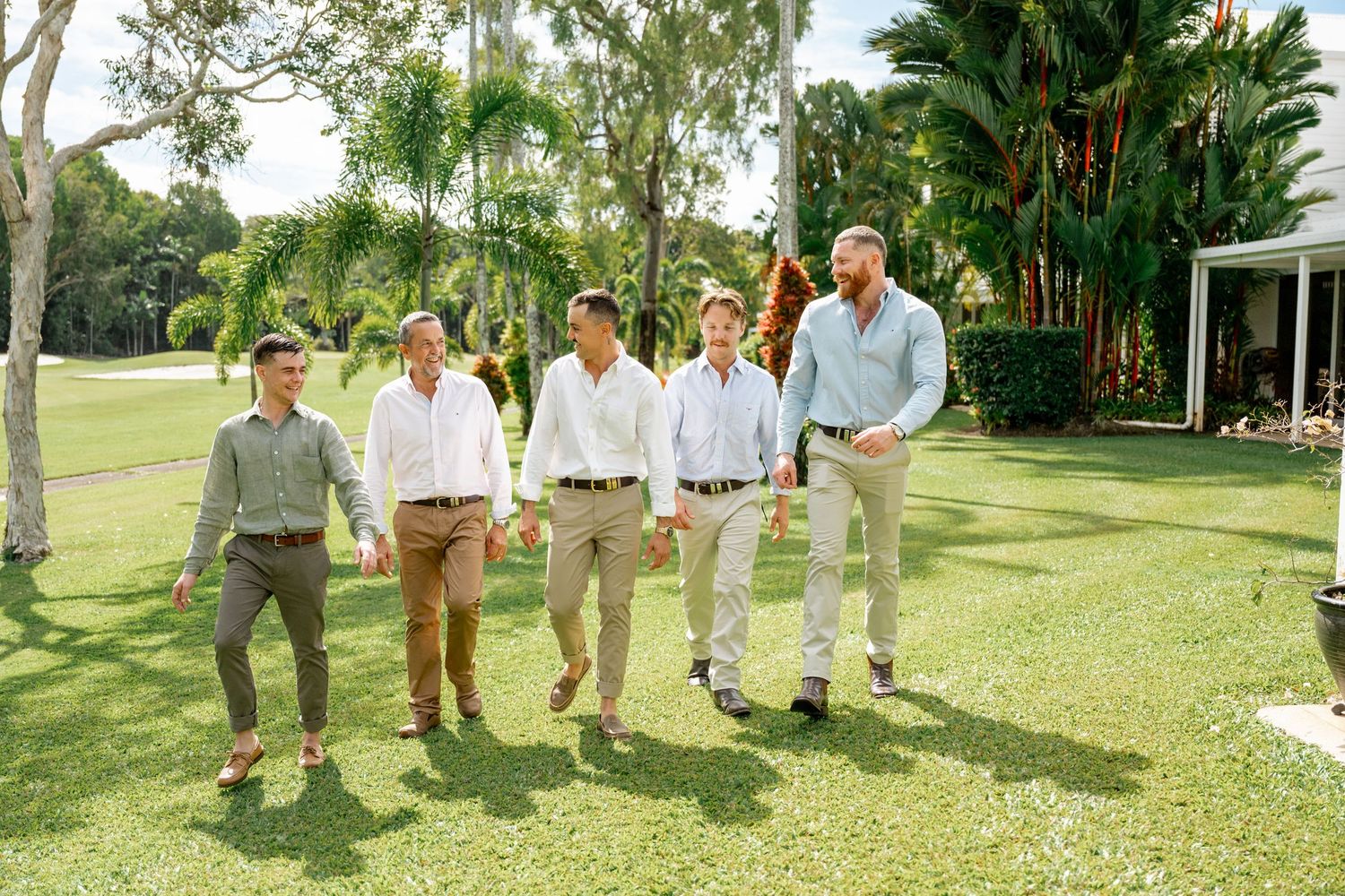 Five groomsmen in smart casual outfits strolling together on green grass with tropical garden backdrop.