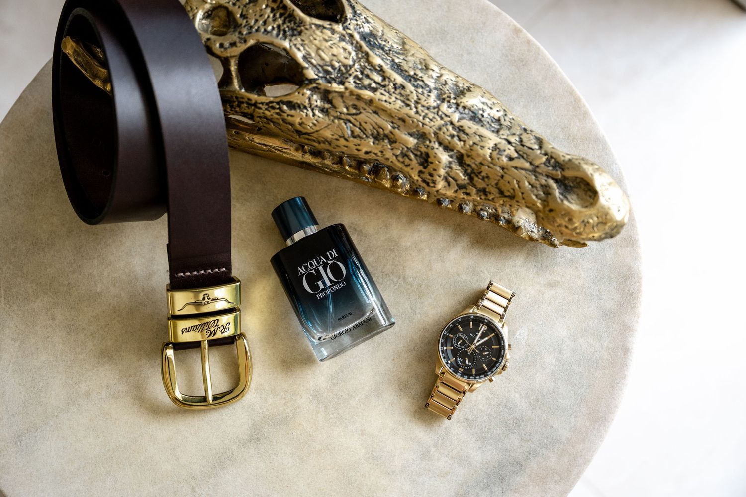 Flat lay of brown leather belt, Acqua di Gio cologne, and gold chronograph watch on marble surface.