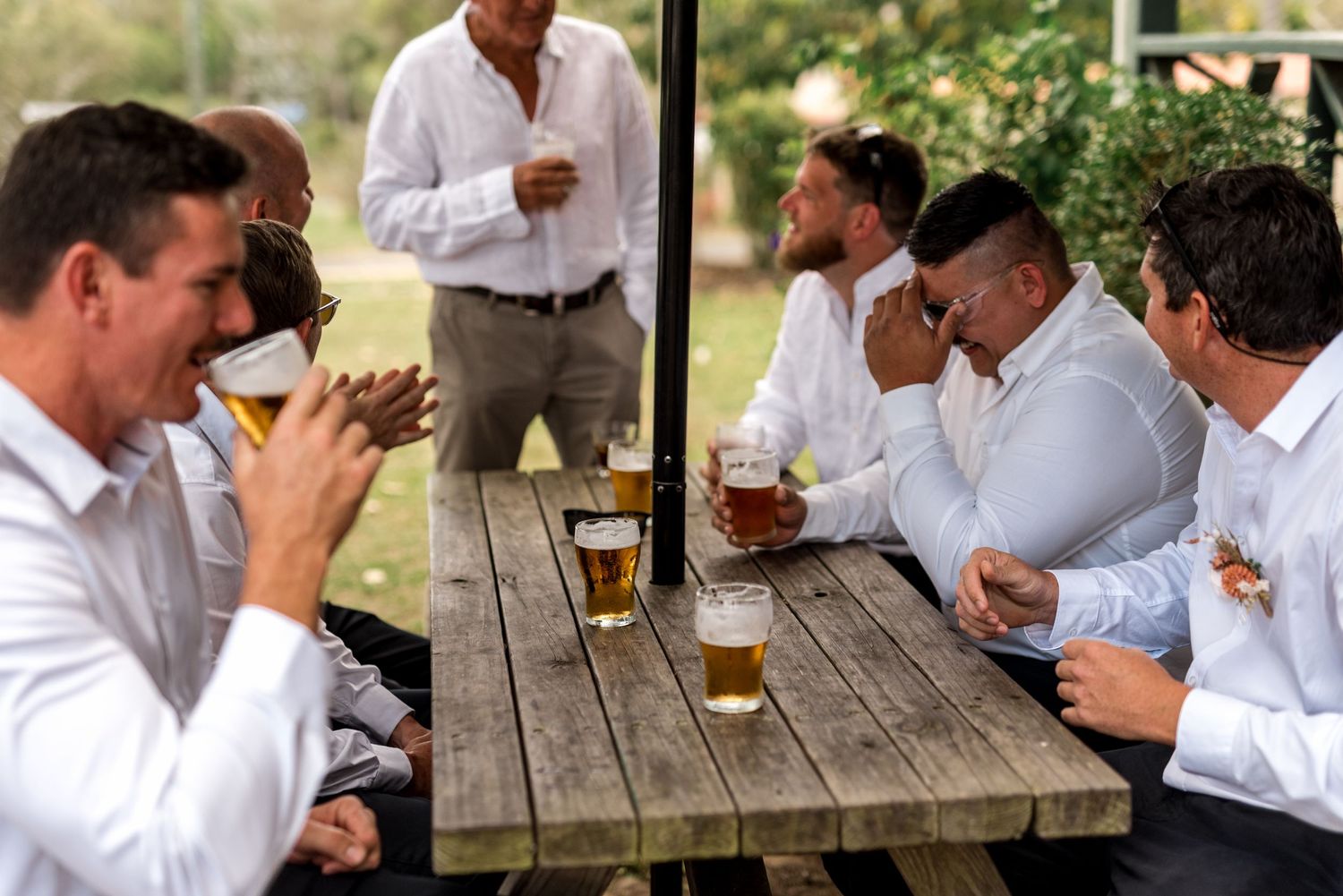 Group of groomsmen in white shirts laughing and drinking beers together at a wooden picnic table outdoors.