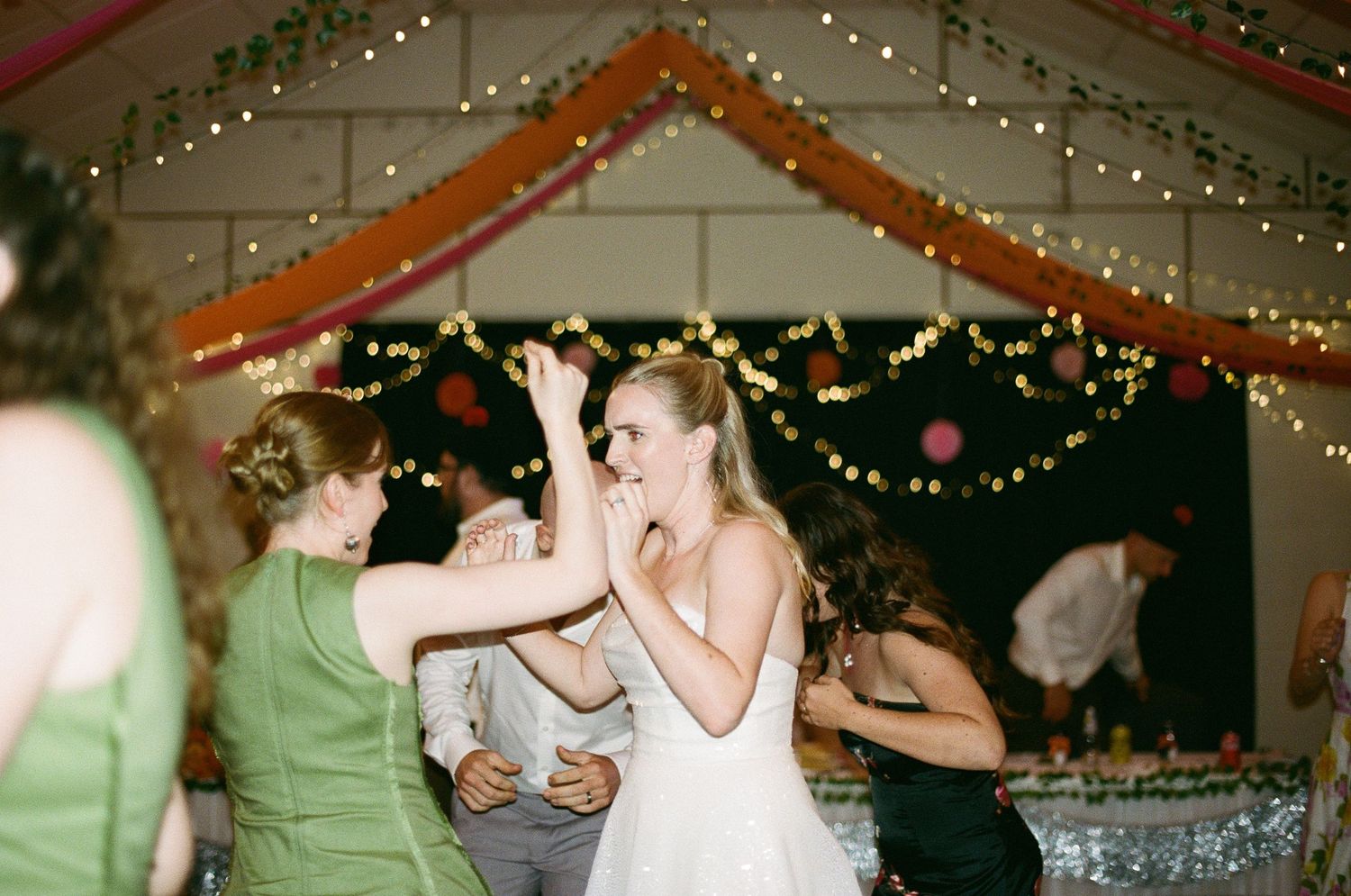 Bride in white gown dancing joyfully with guests under string lights at a wedding reception.