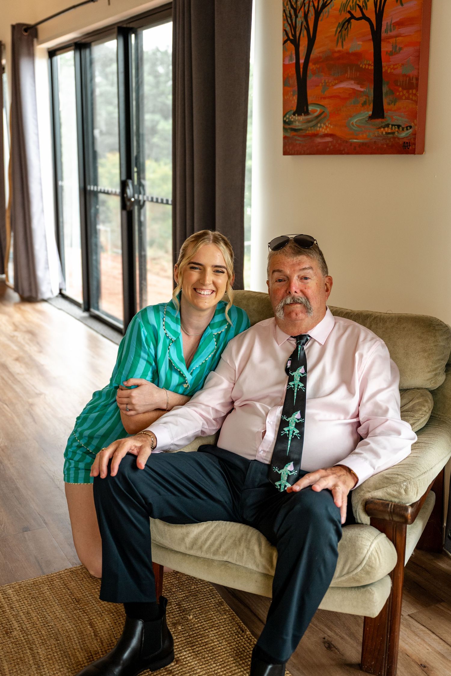 Smiling bride in teal robe posing with older man in pink shirt and novelty tie in armchair.