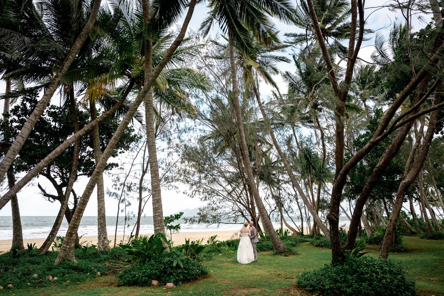 Bride in white wedding gown standing among leaning palm trees on a tropical beach with ocean in background.