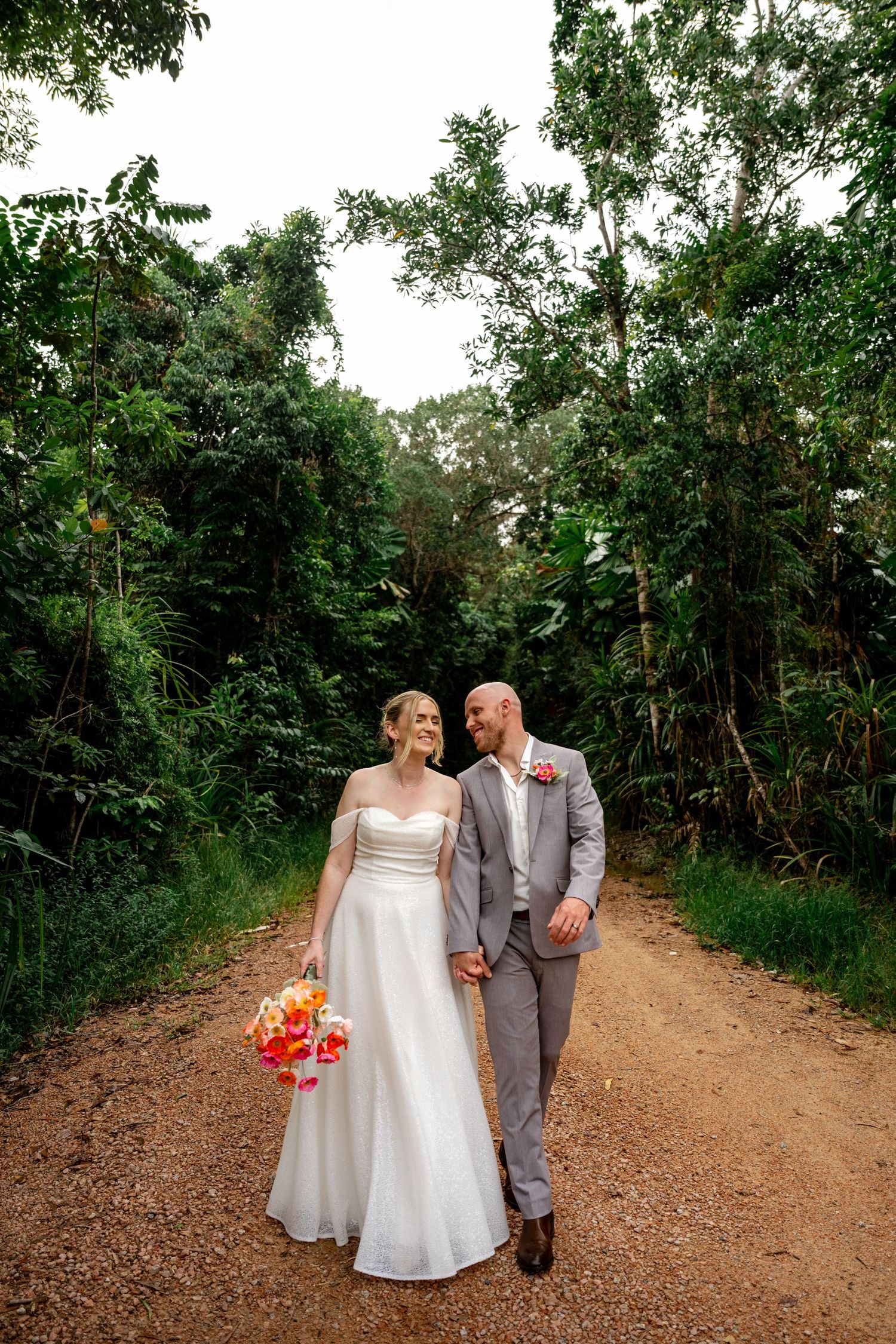 Bride and groom walk hand-in-hand down a lush tropical dirt path, bride holding vibrant orange bouquet.