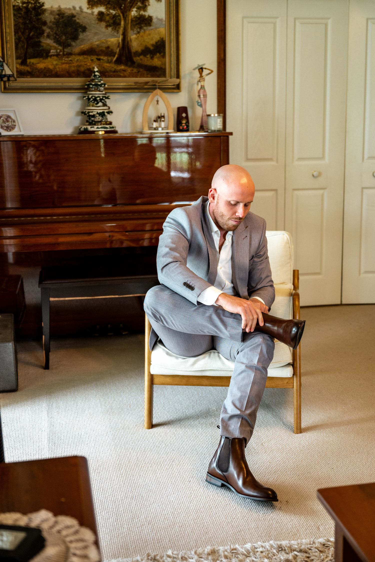 Bald groom in grey suit putting on brown Chelsea boots while seated in armchair before wedding.