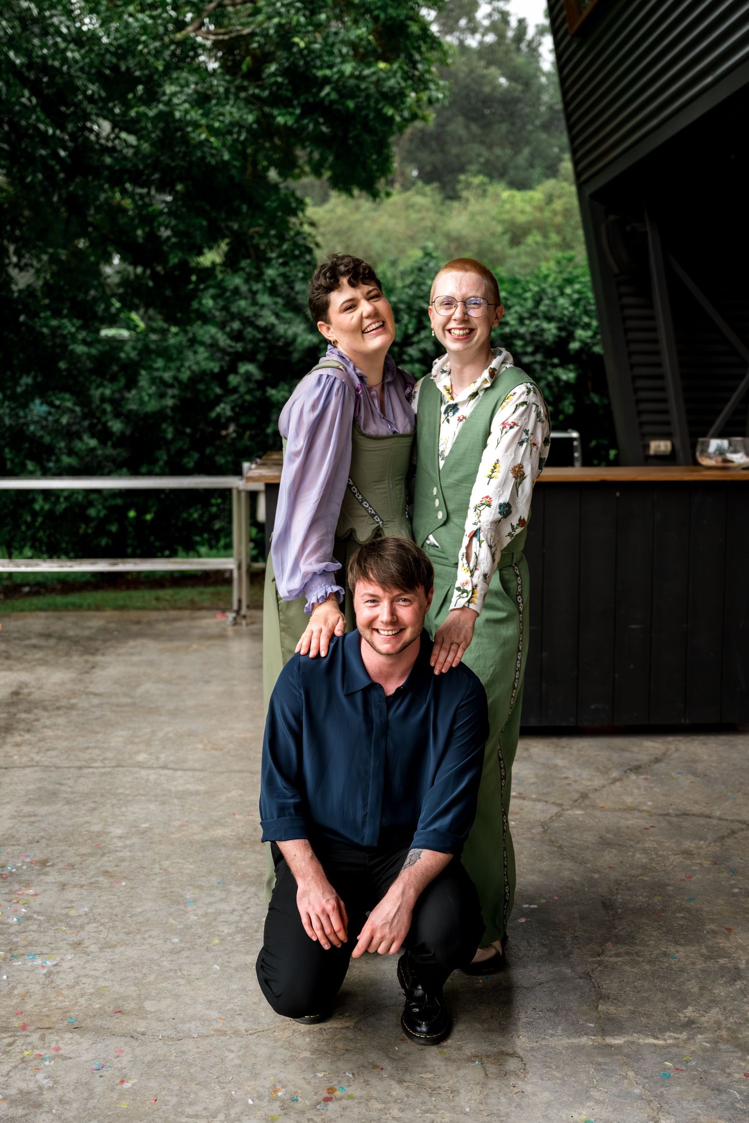 Two brides in green wedding attire posing with a kneeling guest on an outdoor patio.