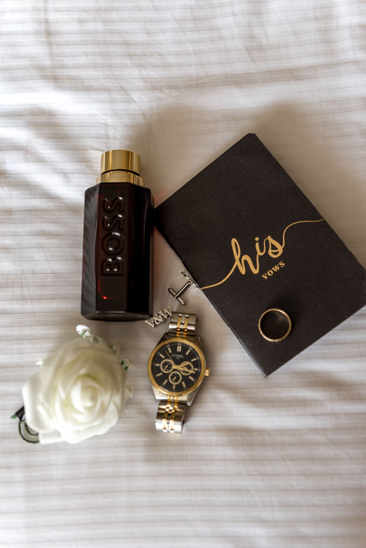 Groom's wedding flat lay featuring Hugo Boss cologne, gold watch, vow book, wedding ring, and white rose on white linen.