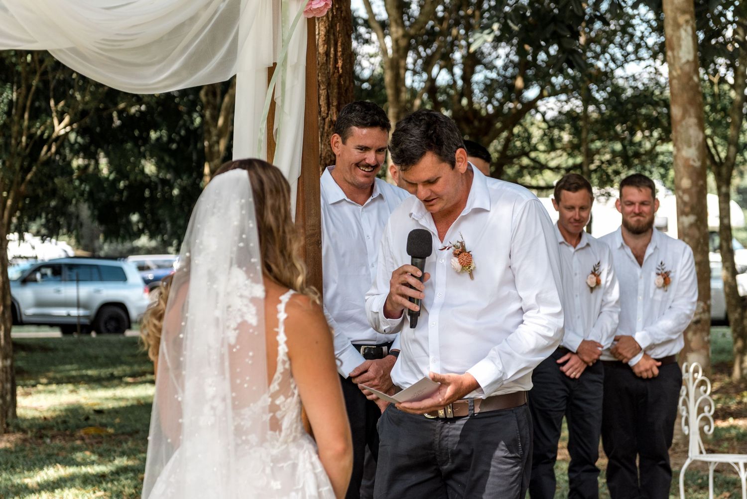 Groom reads vows from card at outdoor wedding with smiling groomsmen standing behind him.