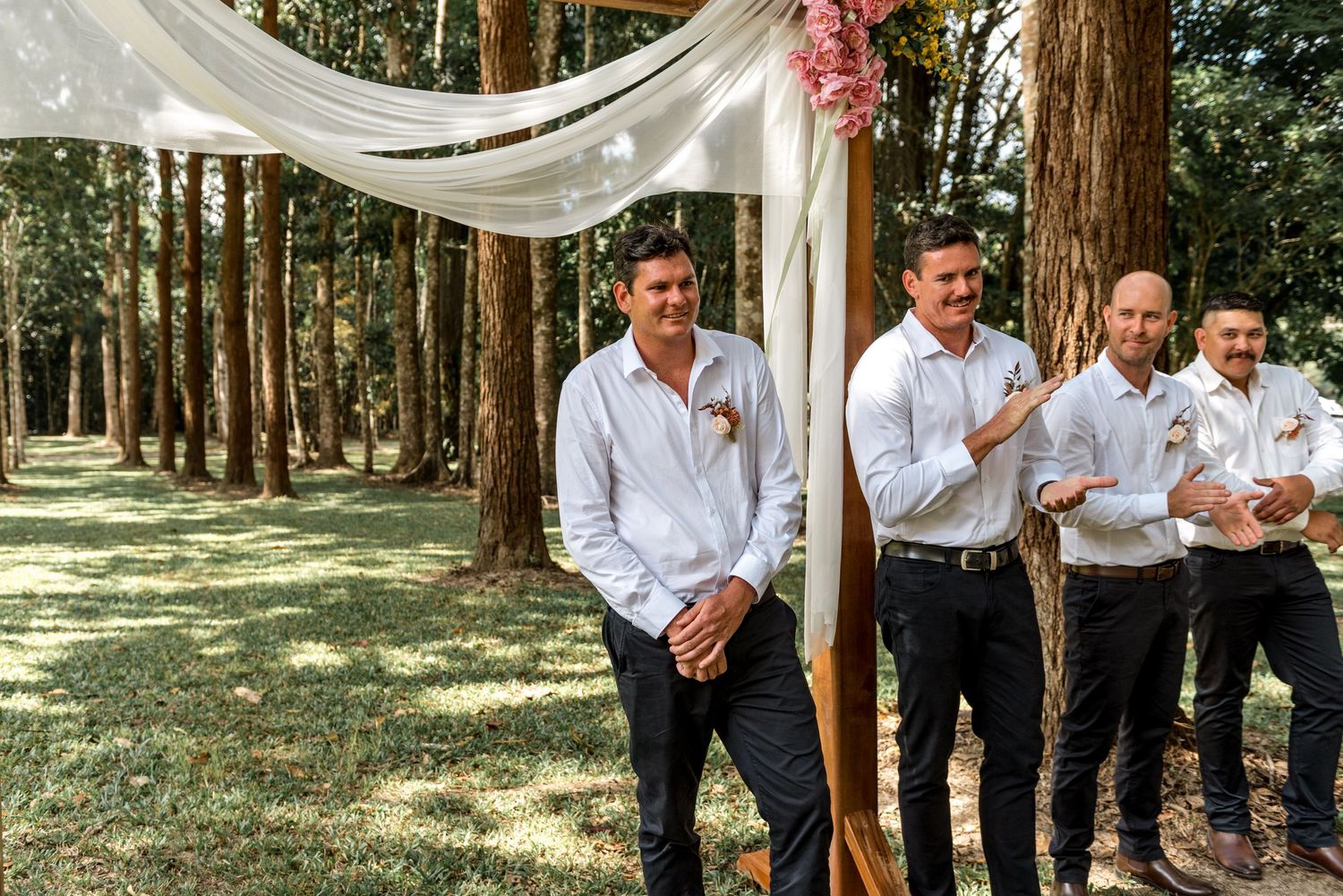 Groom and groomsmen in white shirts smiling under a draped wooden arch in a forest wedding.