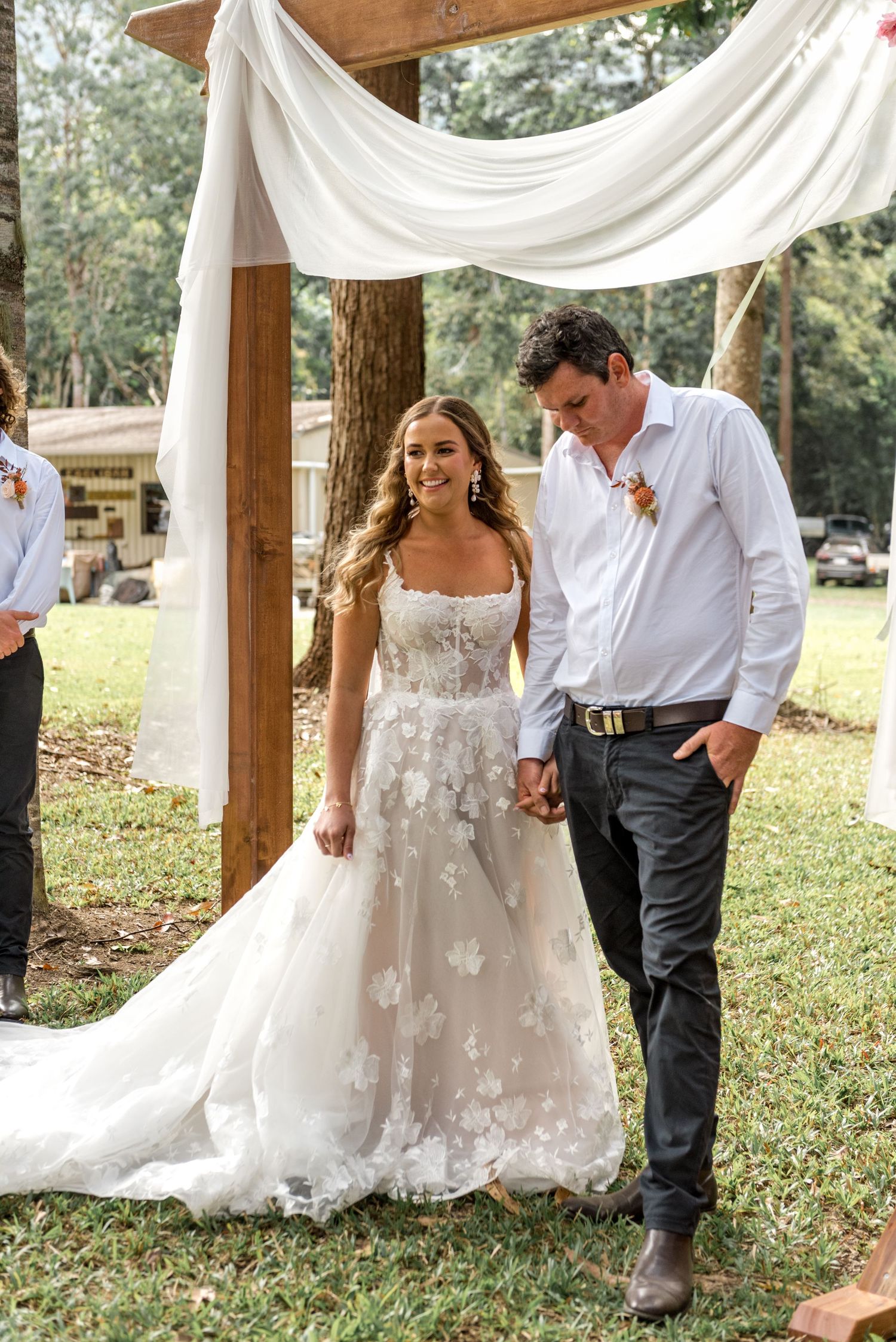 Bride and groom walk together under wooden arch draped with white fabric at outdoor wedding ceremony.