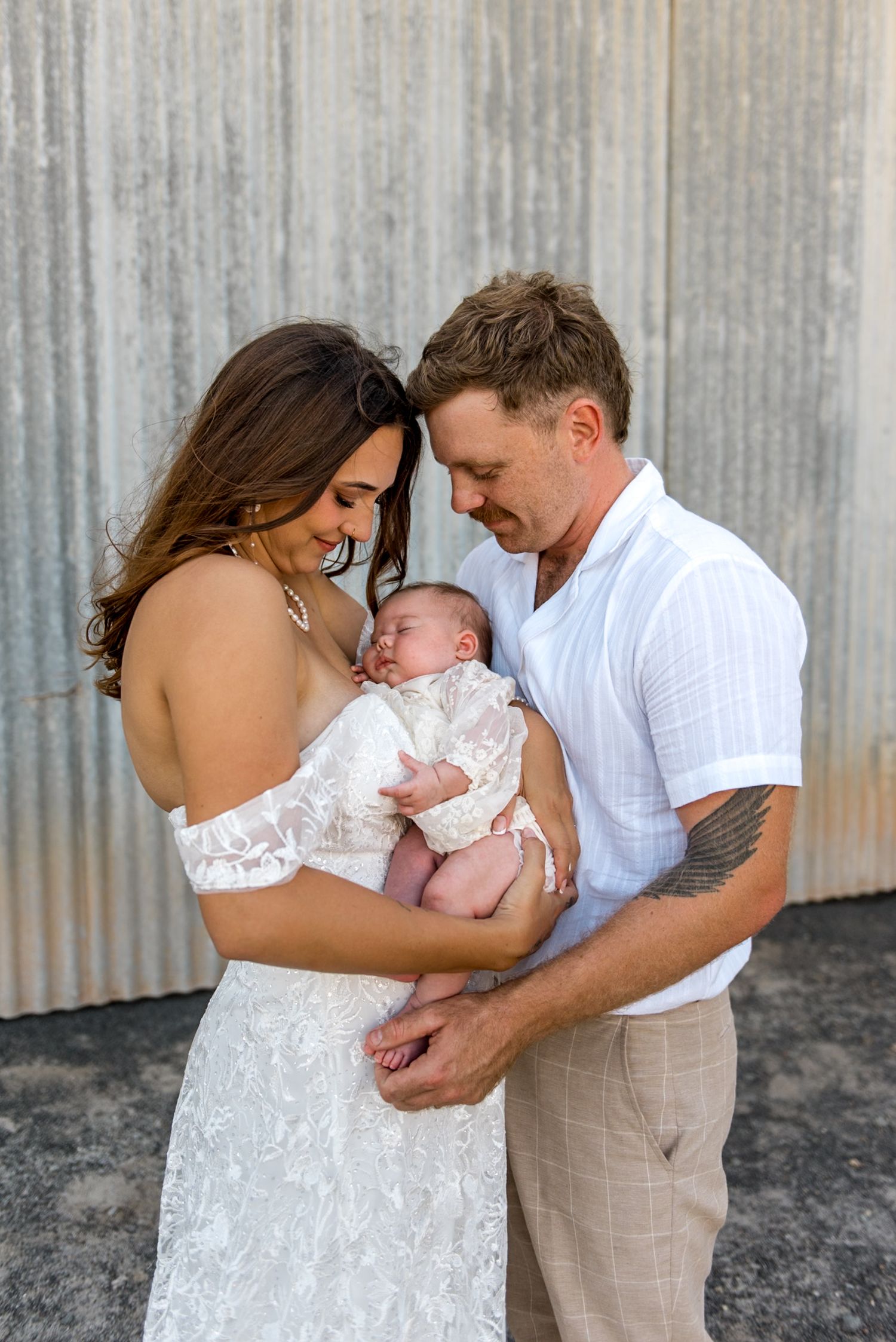 Smiling bride in lace gown and tattooed groom in white shirt gazing lovingly at newborn baby.