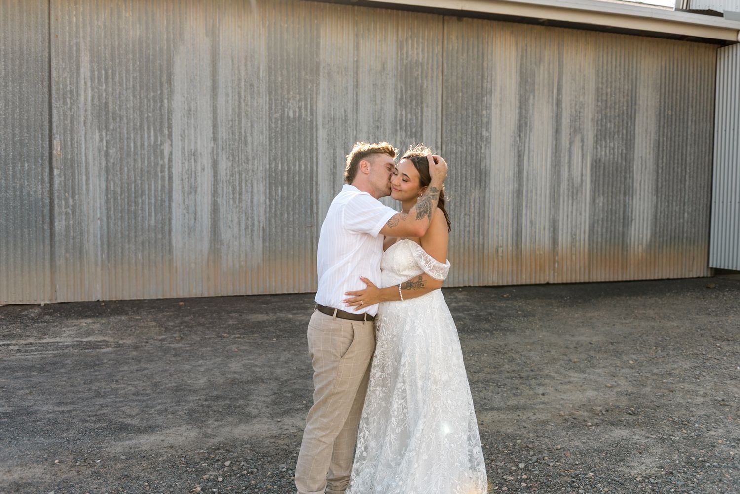 Groom embraces bride in lace off-shoulder gown for a kiss outdoors near a rustic corrugated metal building.