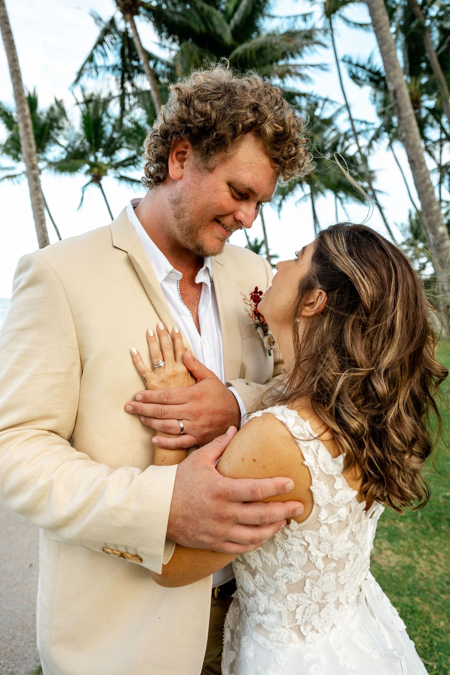 Bride and groom gazing lovingly at each other while embracing beneath tall palm trees on their wedding day.