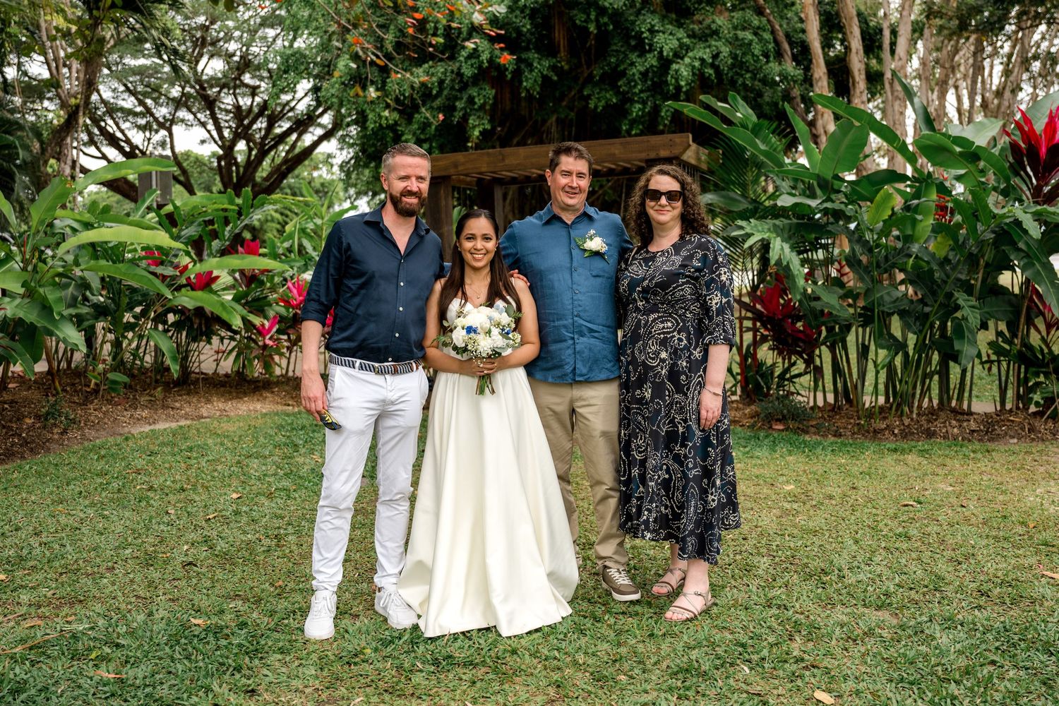 Bride and groom pose with two guests in lush tropical garden with red flowers after wedding at Little Cove, Port Douglas.