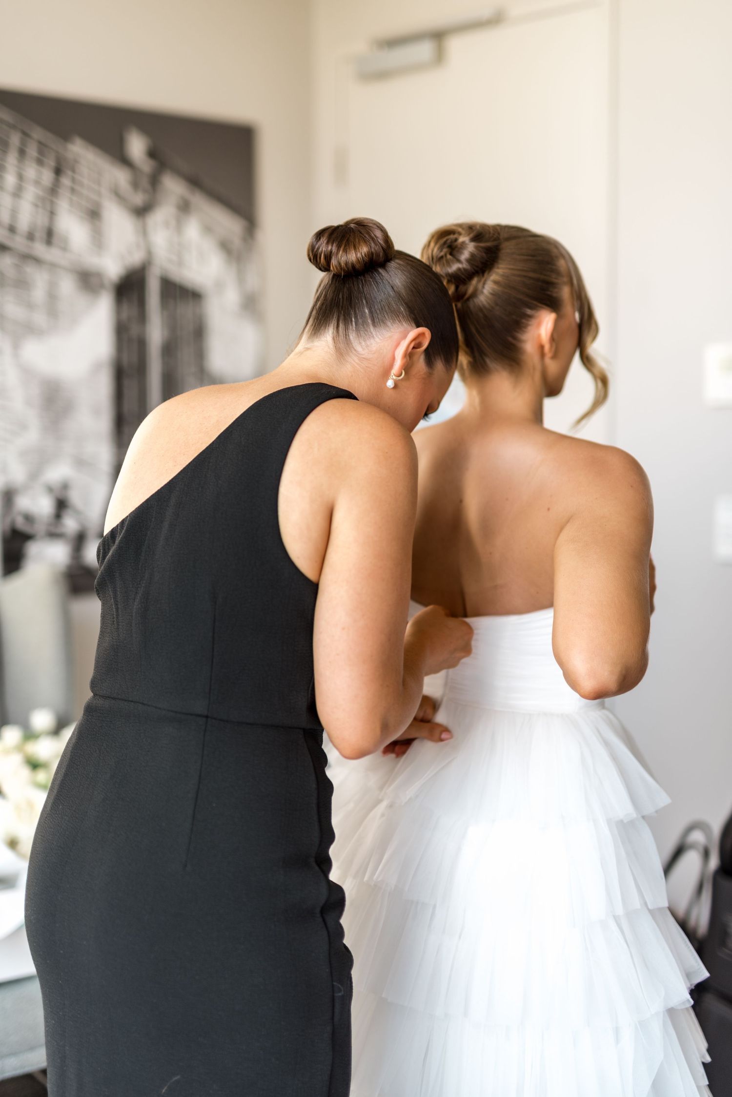 Bridesmaid in black dress helping bride fasten strapless layered white wedding gown from behind.