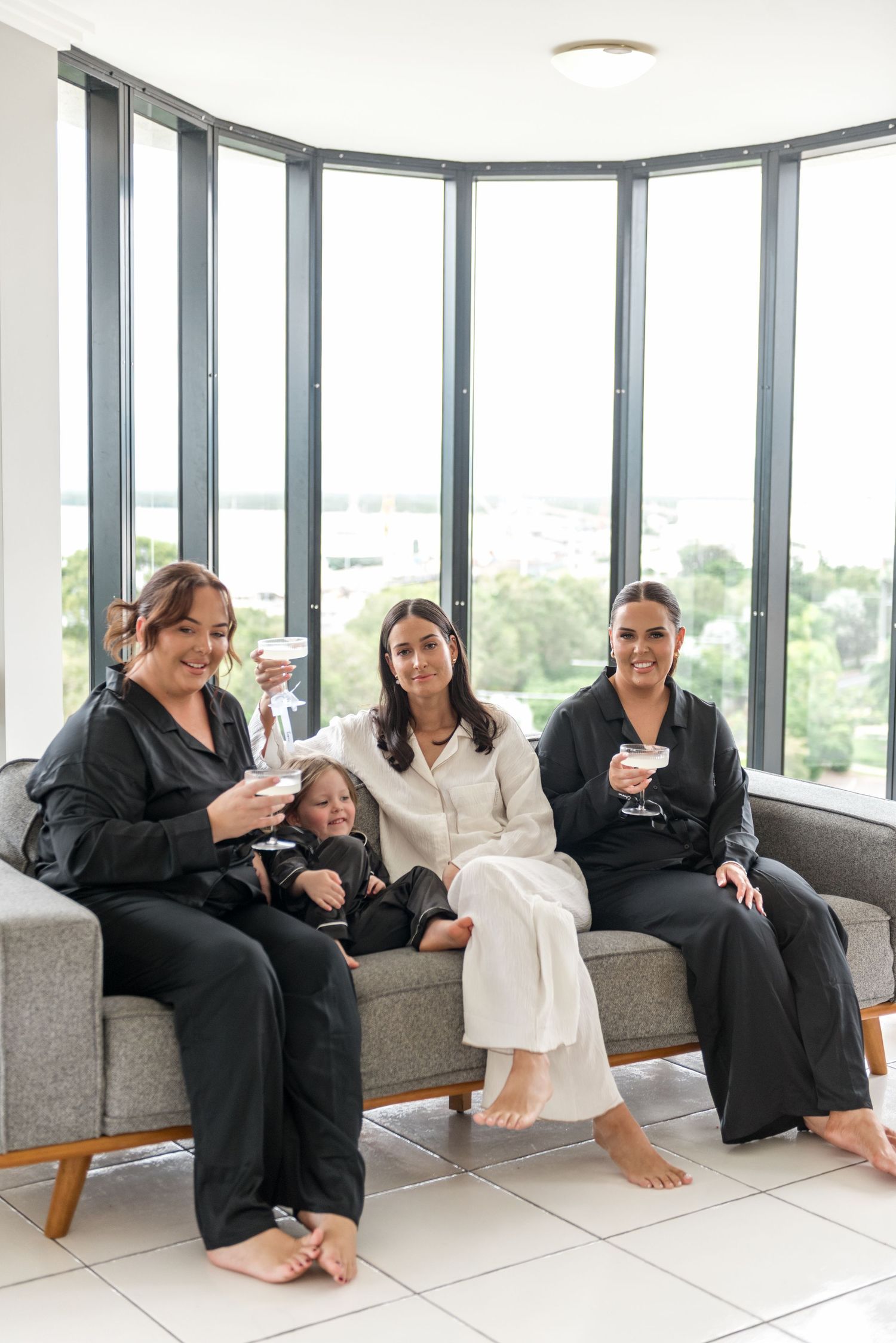 Four women including a child in matching black satin pajamas smile and raise glasses on a grey sofa by panoramic windows.