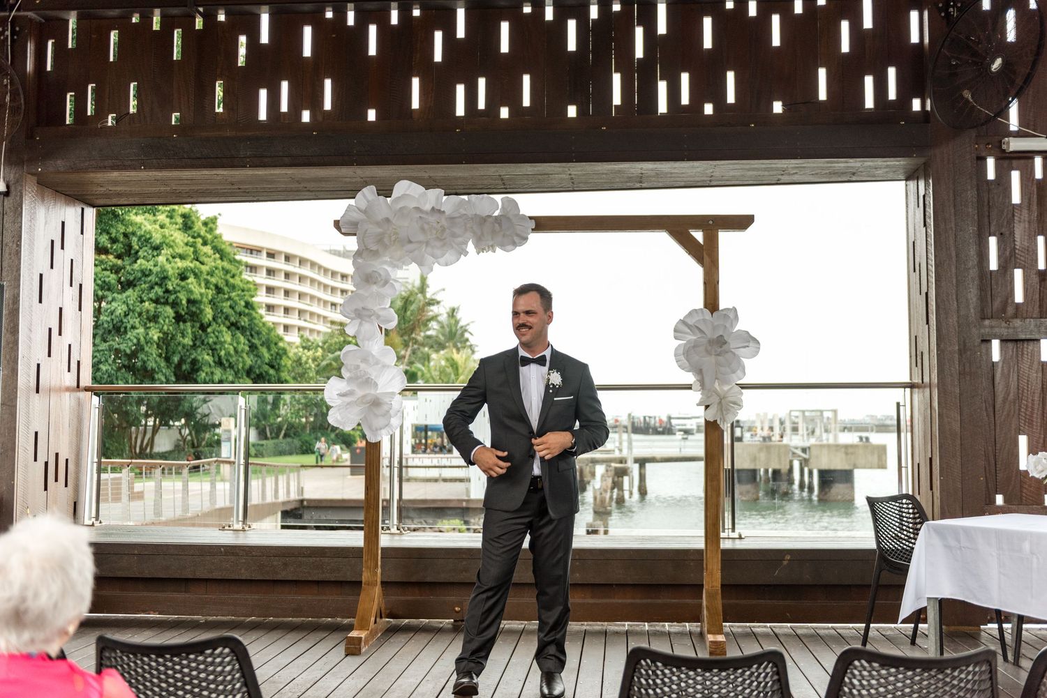 Groom in black tuxedo standing at a waterfront wedding ceremony altar with white floral arrangements.
