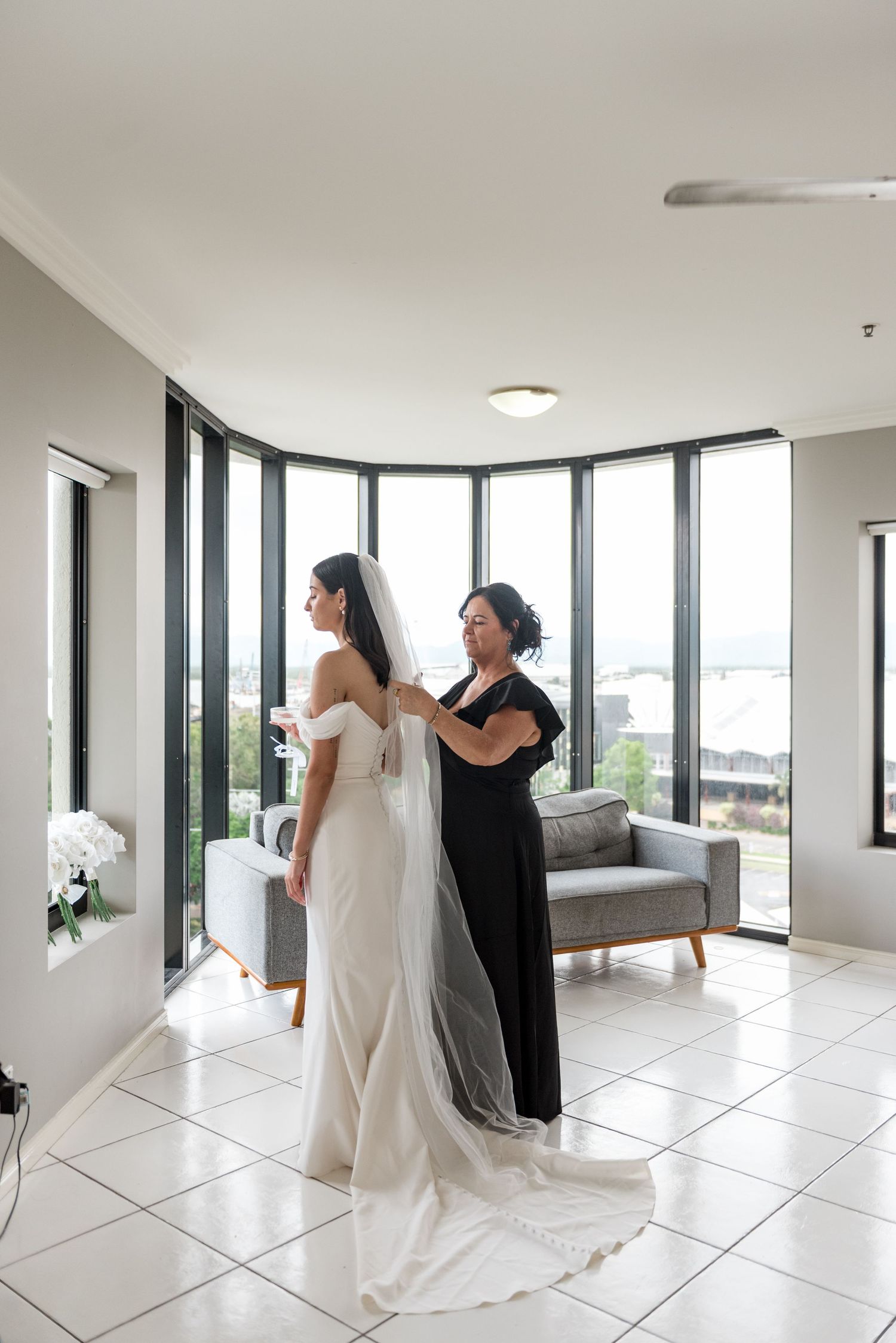 Mother of the bride in black dress attaches veil to bride's elegant white wedding gown by bay windows.