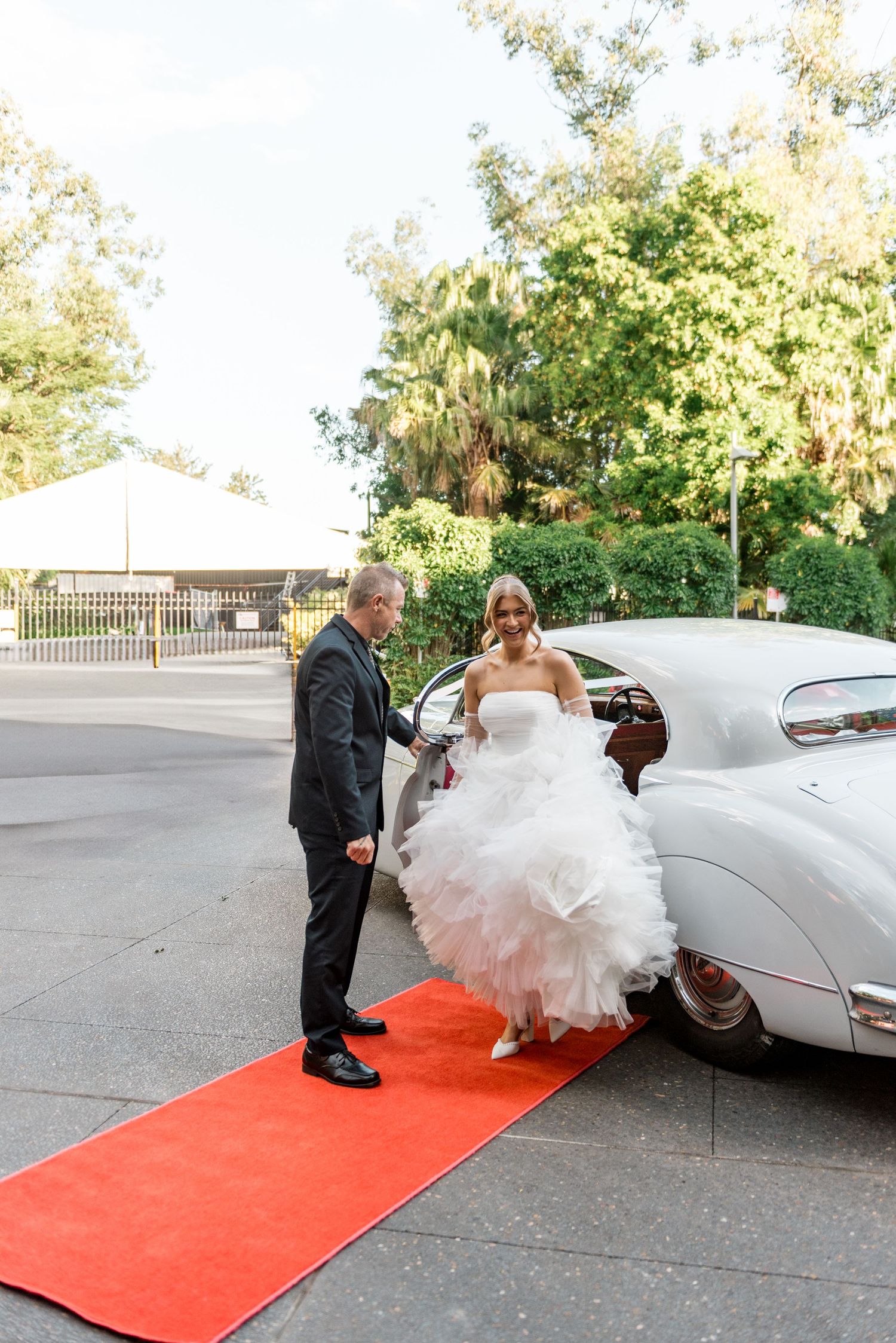 Bride in tiered white dress exits vintage white car on red carpet with escort.