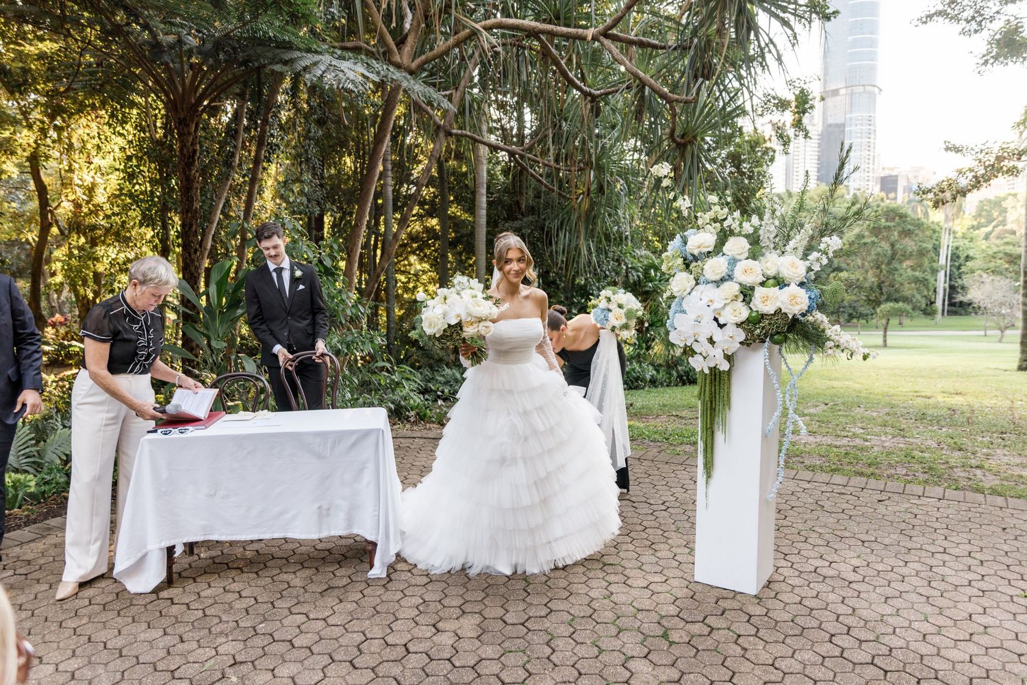 Bride signing wedding register at outdoor ceremony with white floral arrangements nearby.