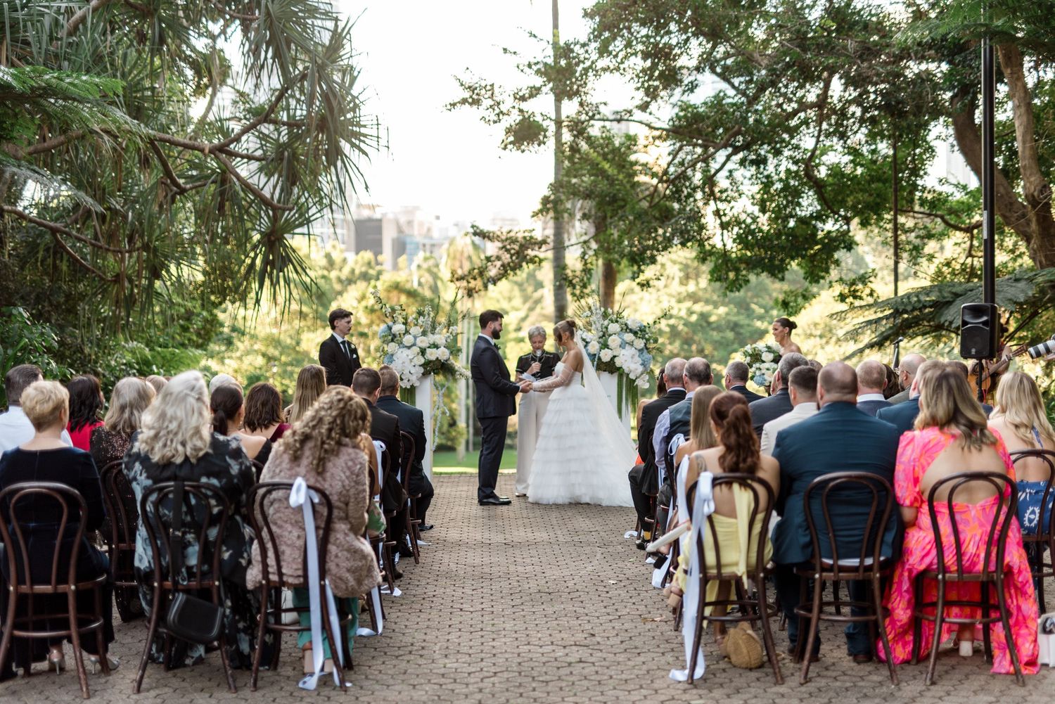 Outdoor wedding ceremony with guests seated on mismatched chairs along a brick path surrounded by lush greenery.