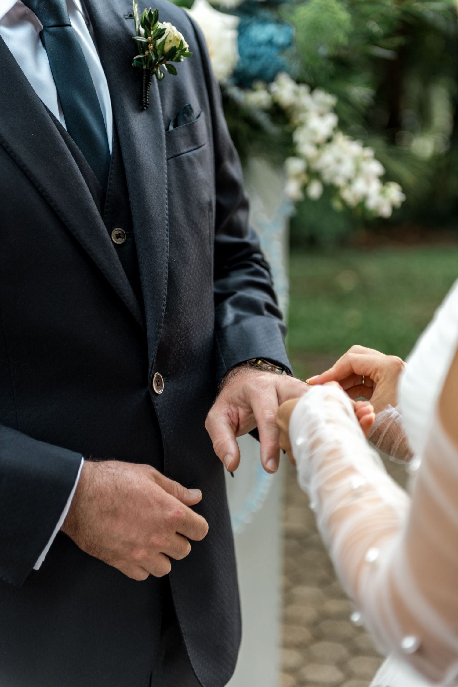 Bride placing wedding ring on groom's finger during outdoor ceremony, groom wearing dark suit with boutonniere.