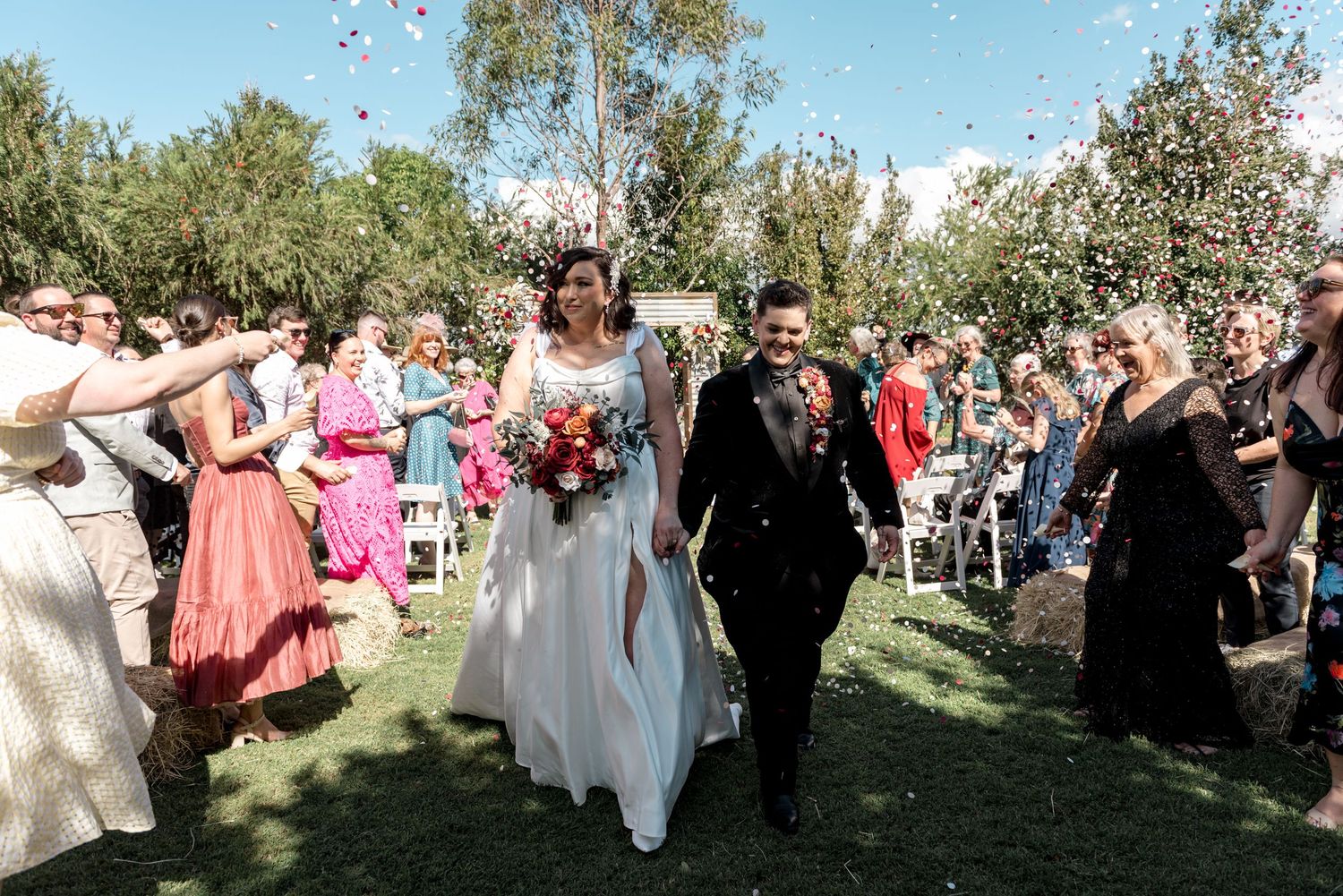 Newlyweds walk down aisle as guests throw colorful confetti at outdoor garden wedding ceremony.
