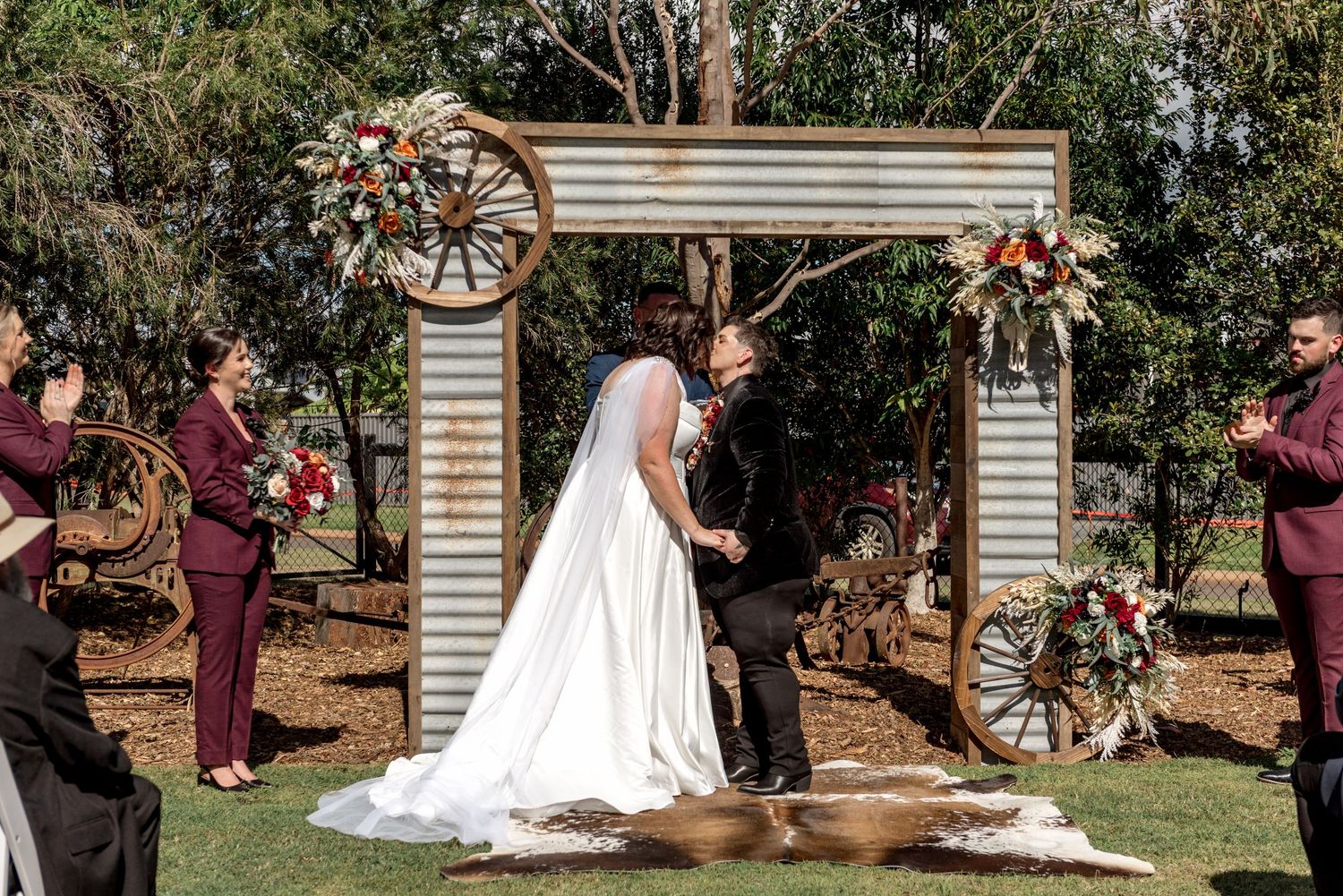 Bride and groom share first kiss under rustic wagon wheel arch at outdoor wedding ceremony.