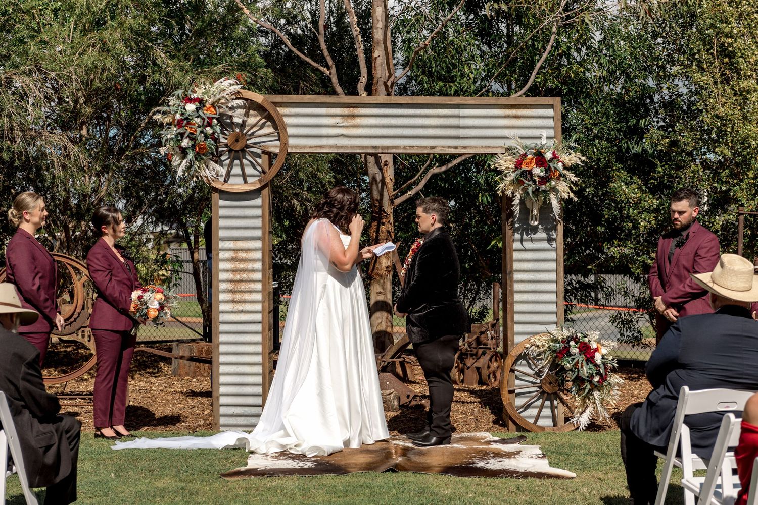 Bride and groom exchange vows at rustic outdoor wedding ceremony beneath a corrugated iron arch with wagon wheels.