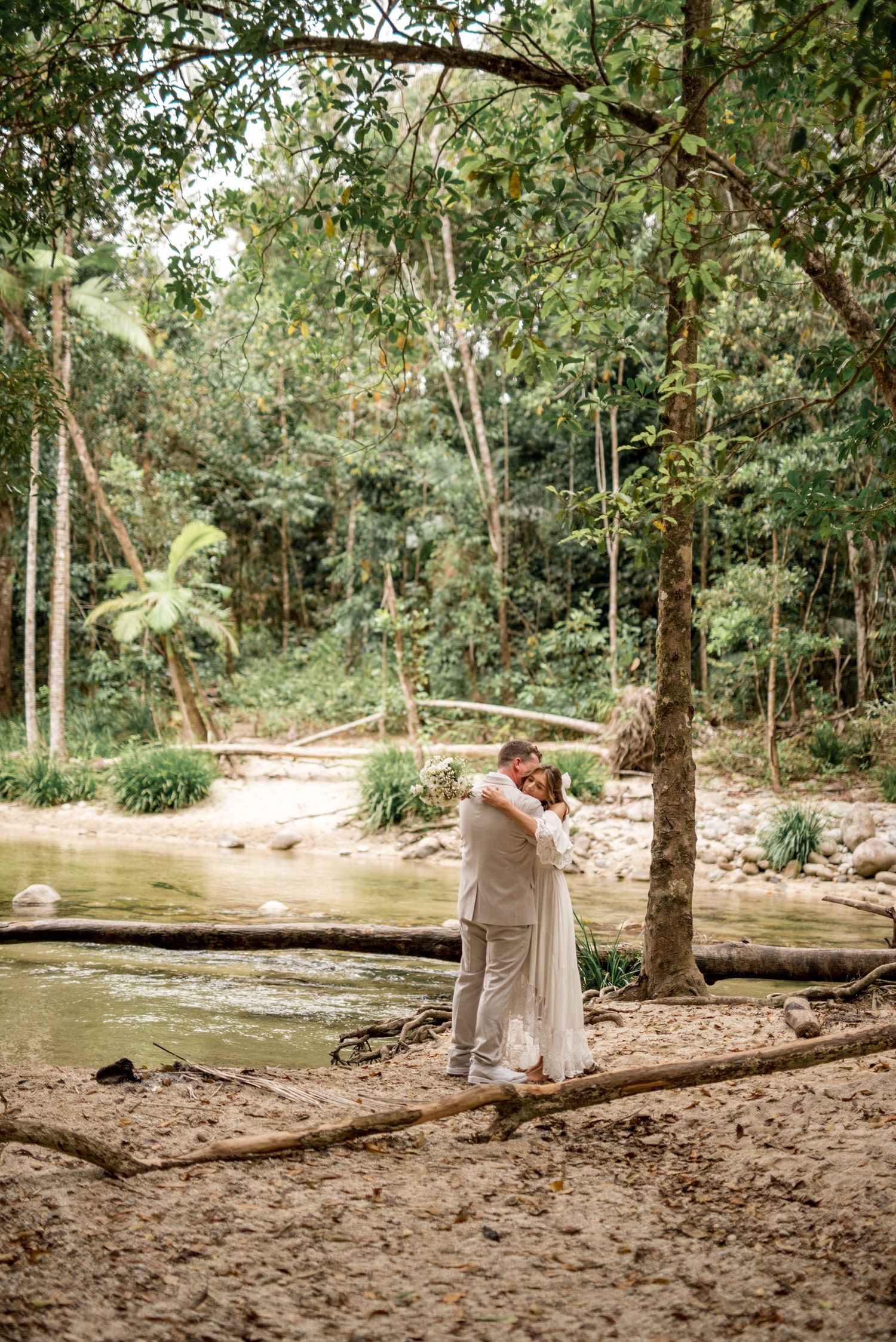 Bride and groom sharing a hug beside a tranquil forest creek, surrounded by lush tropical greenery.