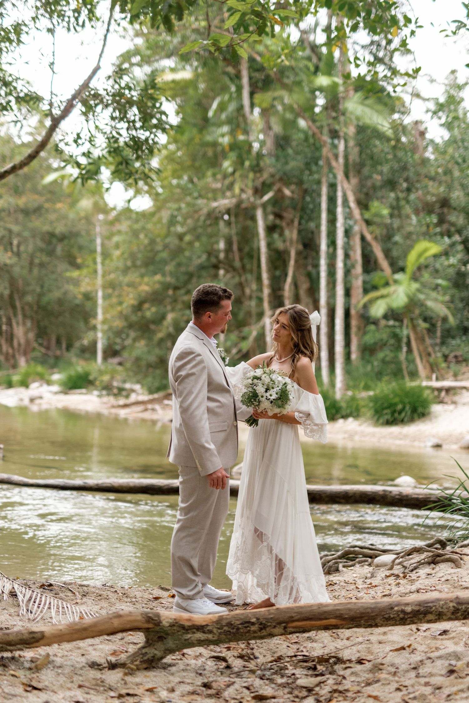Bride and groom stand together on a sandy riverbank surrounded by lush green forest trees.