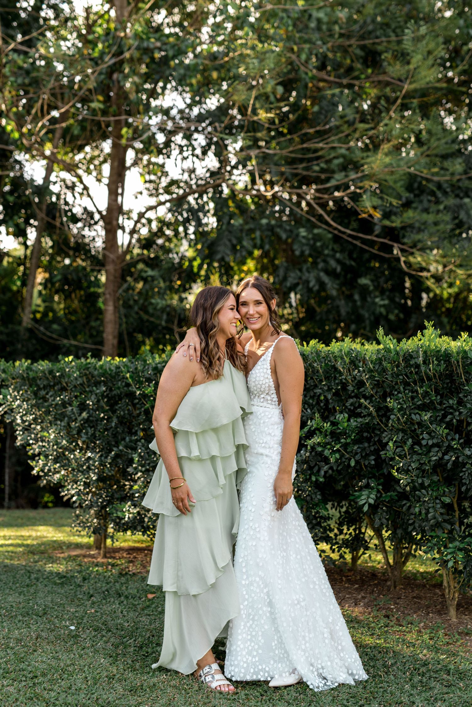 Bride in lace gown and bridesmaid in sage green dress pose together in garden.