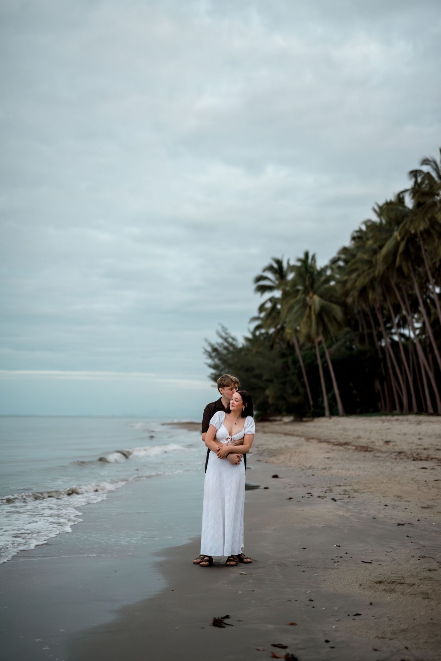 Couple holding each other on a wet sandy beach with palm trees and ocean waves behind them.