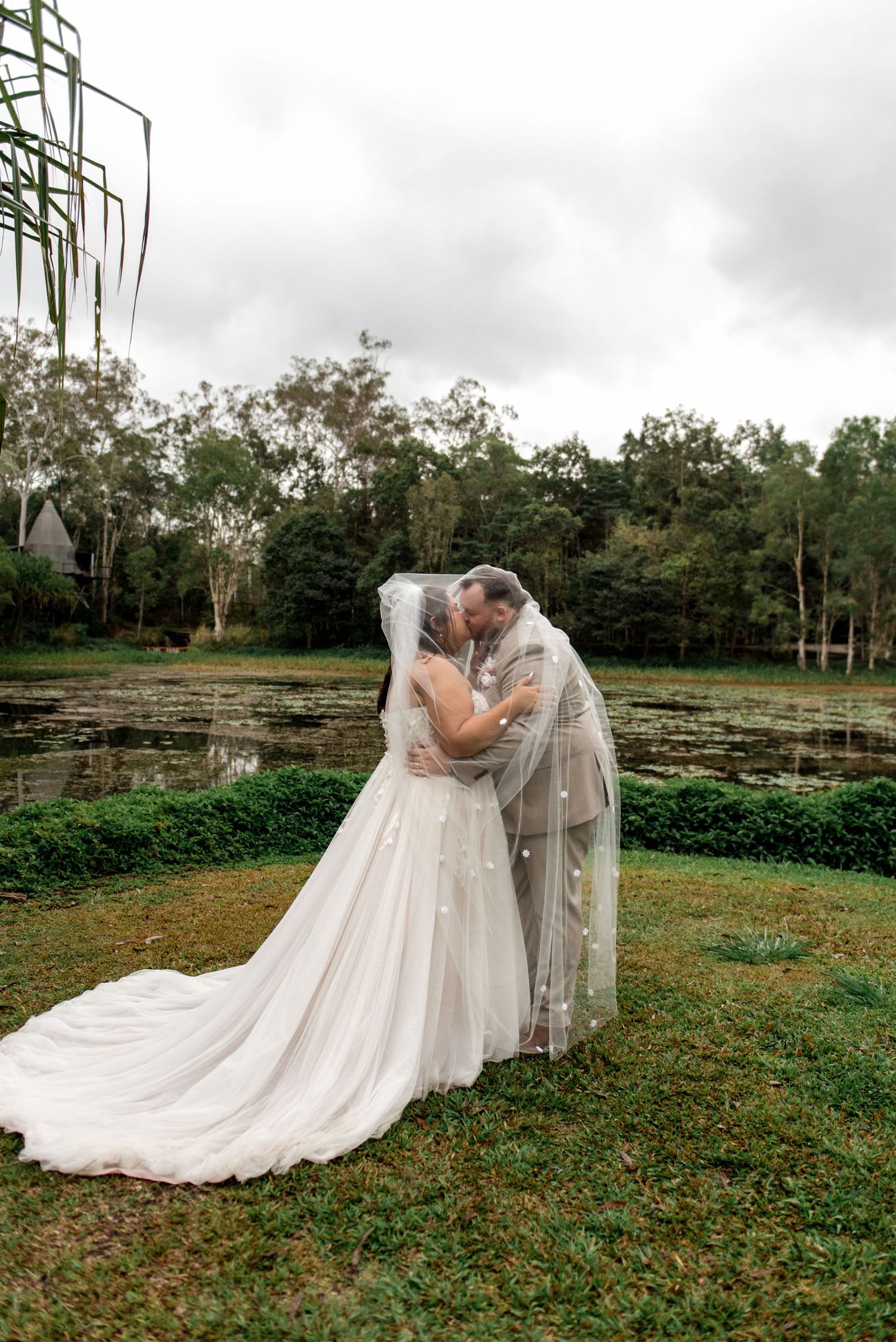 Newlyweds kiss romantically under a pearl-dotted veil beside a tranquil lily pond.