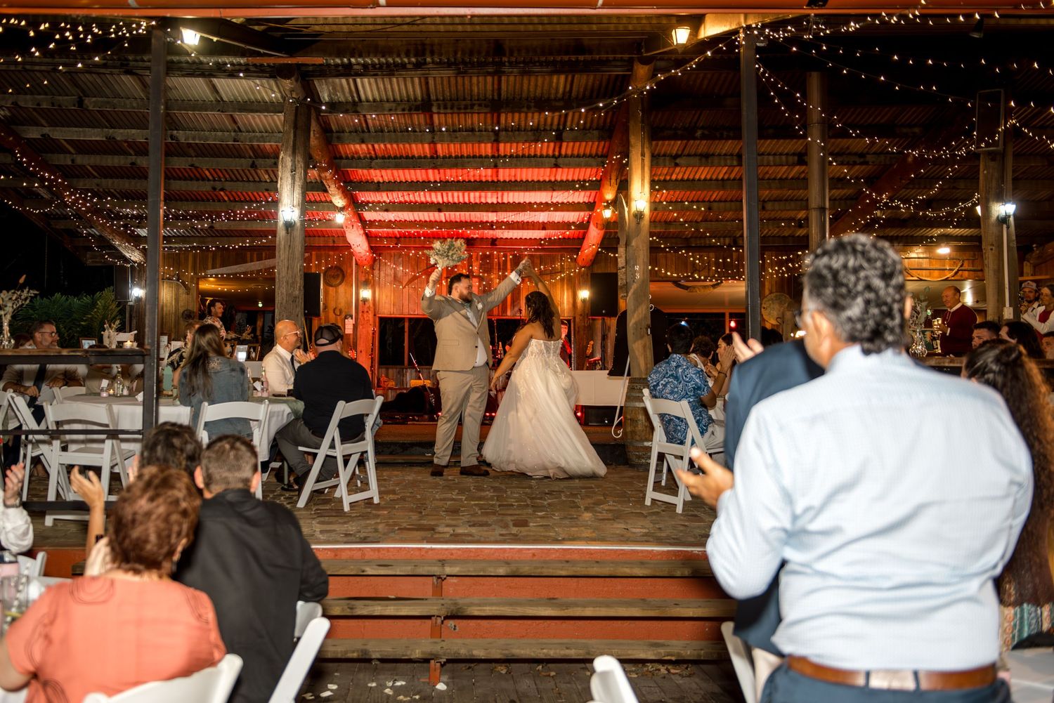 Bride and groom share first dance at rustic barn wedding reception with string lights and seated guests.