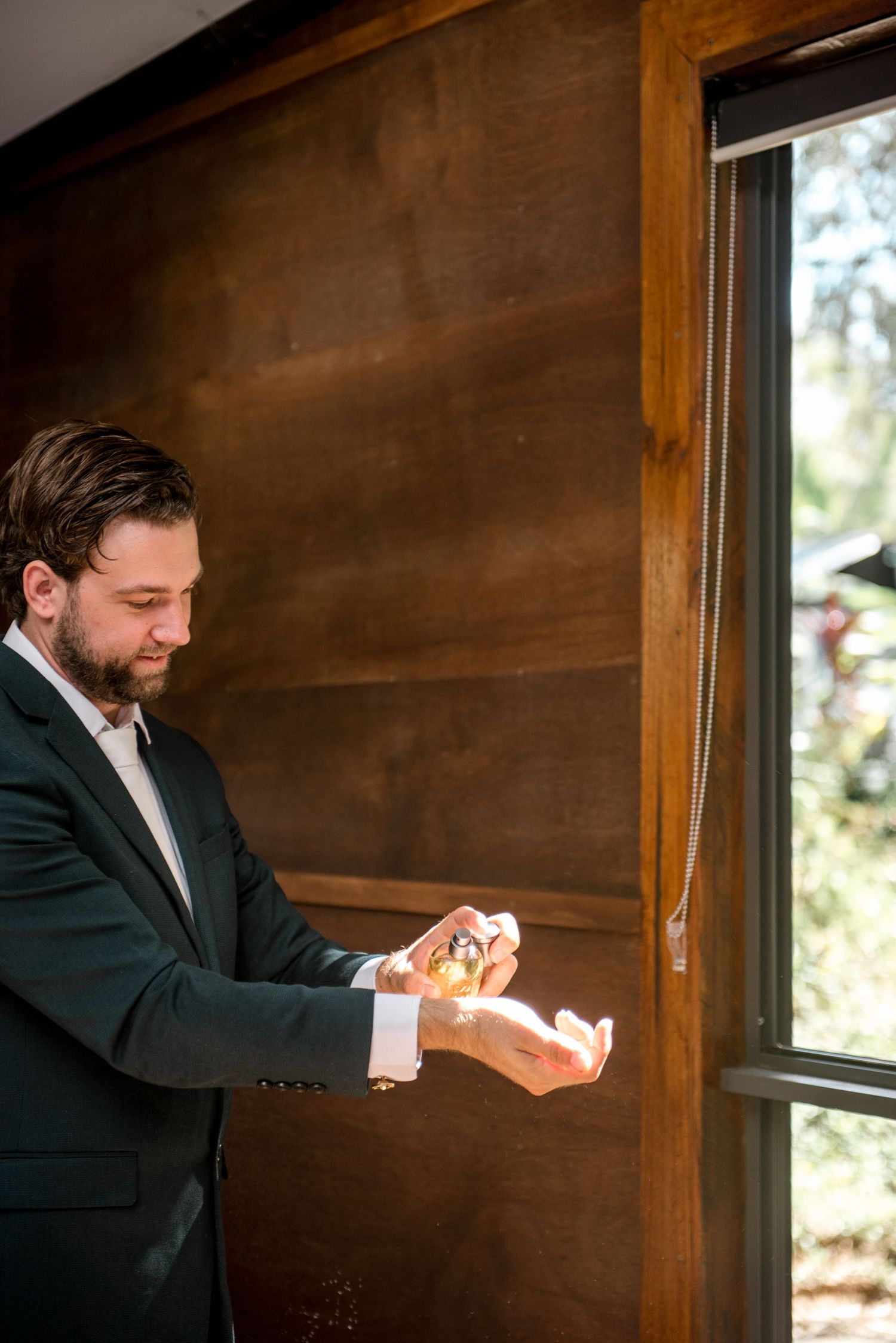 Groom in dark suit applying cologne before wedding ceremony in rustic wooden room.